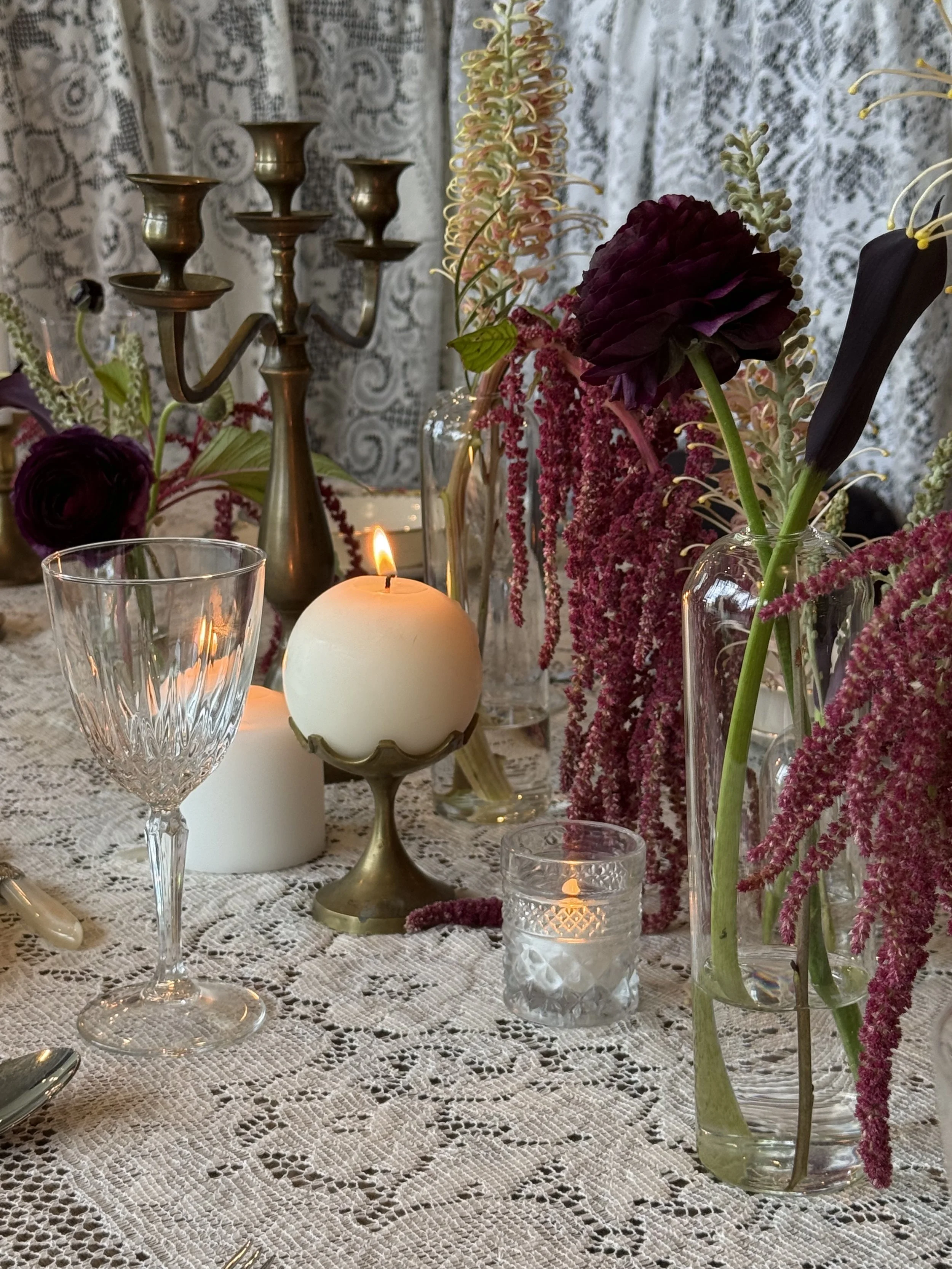 A table decorated with floral arrangements in glass vases, lit candles, a white lace tablecloth, a bronze candelabra, and a backdrop of lace curtains.
