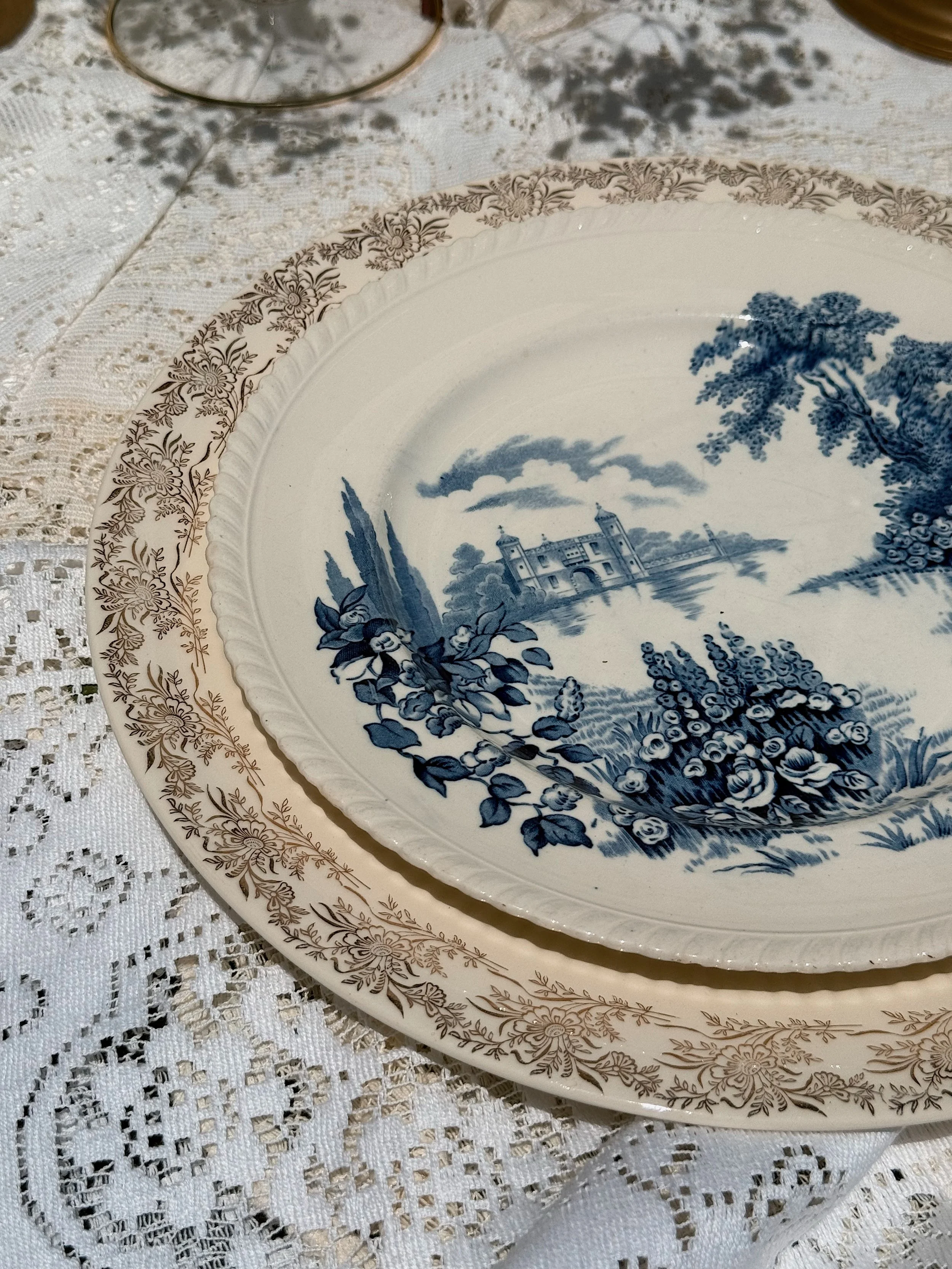 Close-up view of two decorative vintage plates with blue and white landscape scenes and floral patterns, placed on a lace tablecloth.