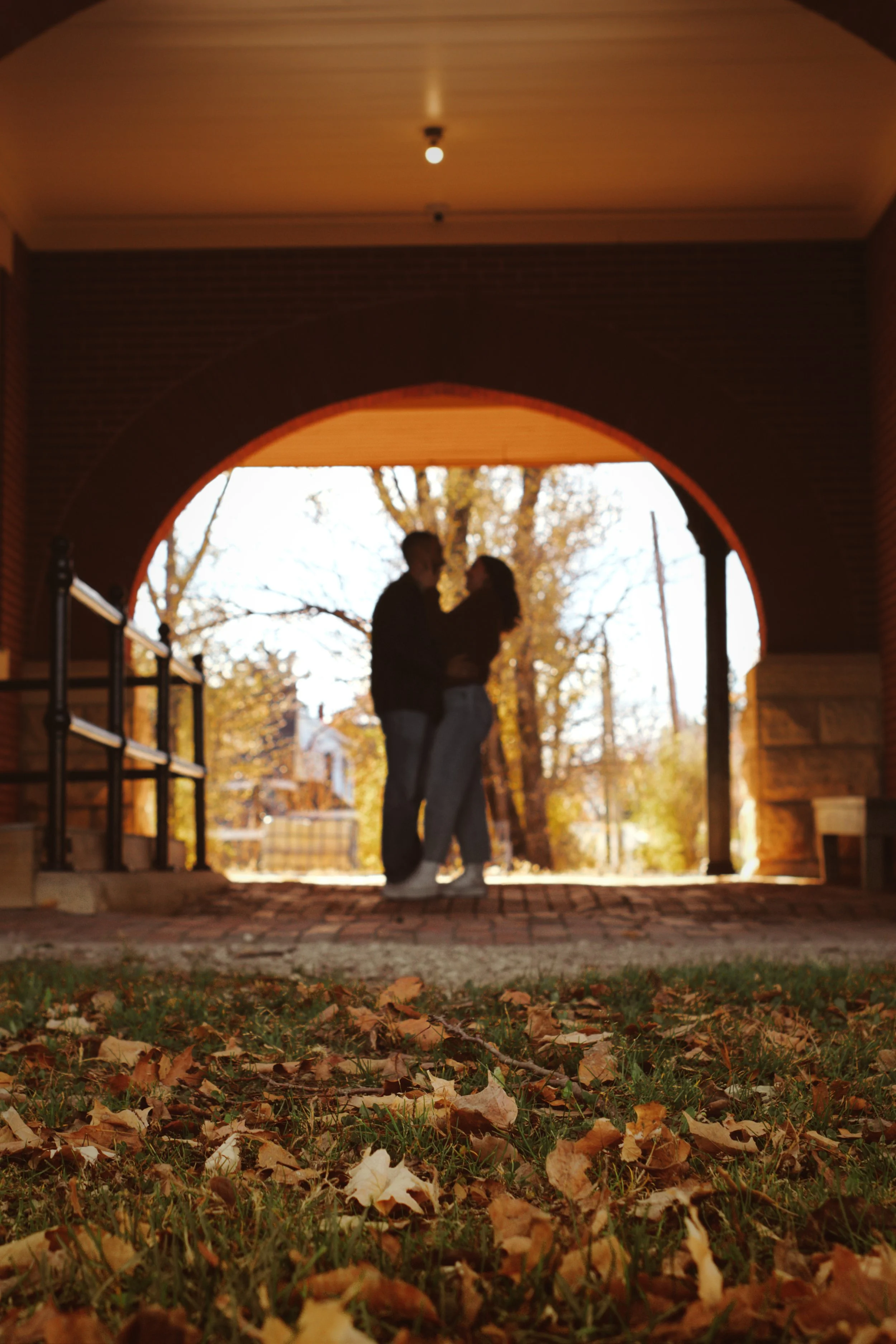 A couple standing closely together under an archway, with autumn trees in the background, viewed from ground level with fallen leaves in the foreground.
