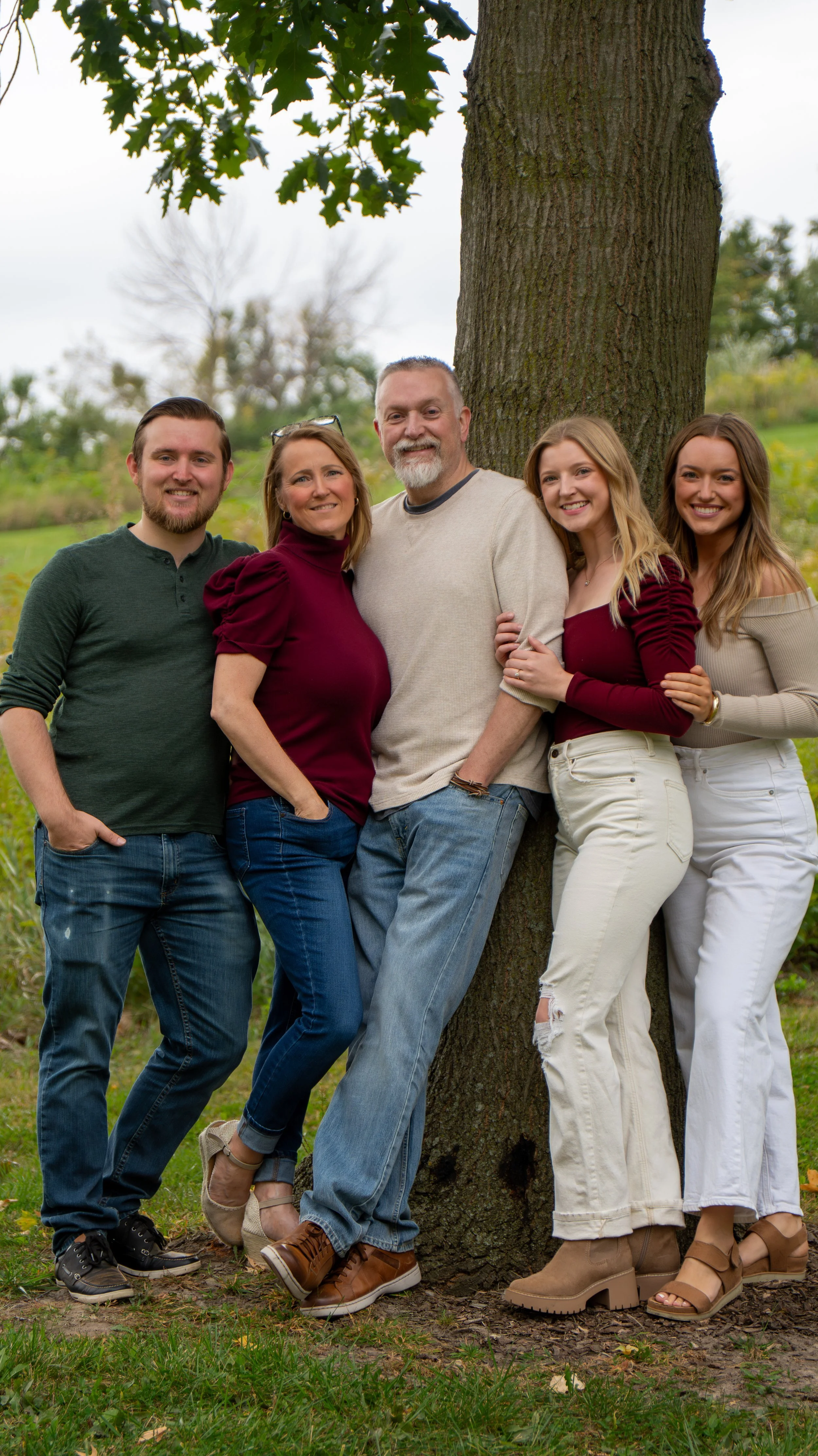 A family of five standing outdoors by a tree in a park, smiling and dressed in casual autumn clothing.