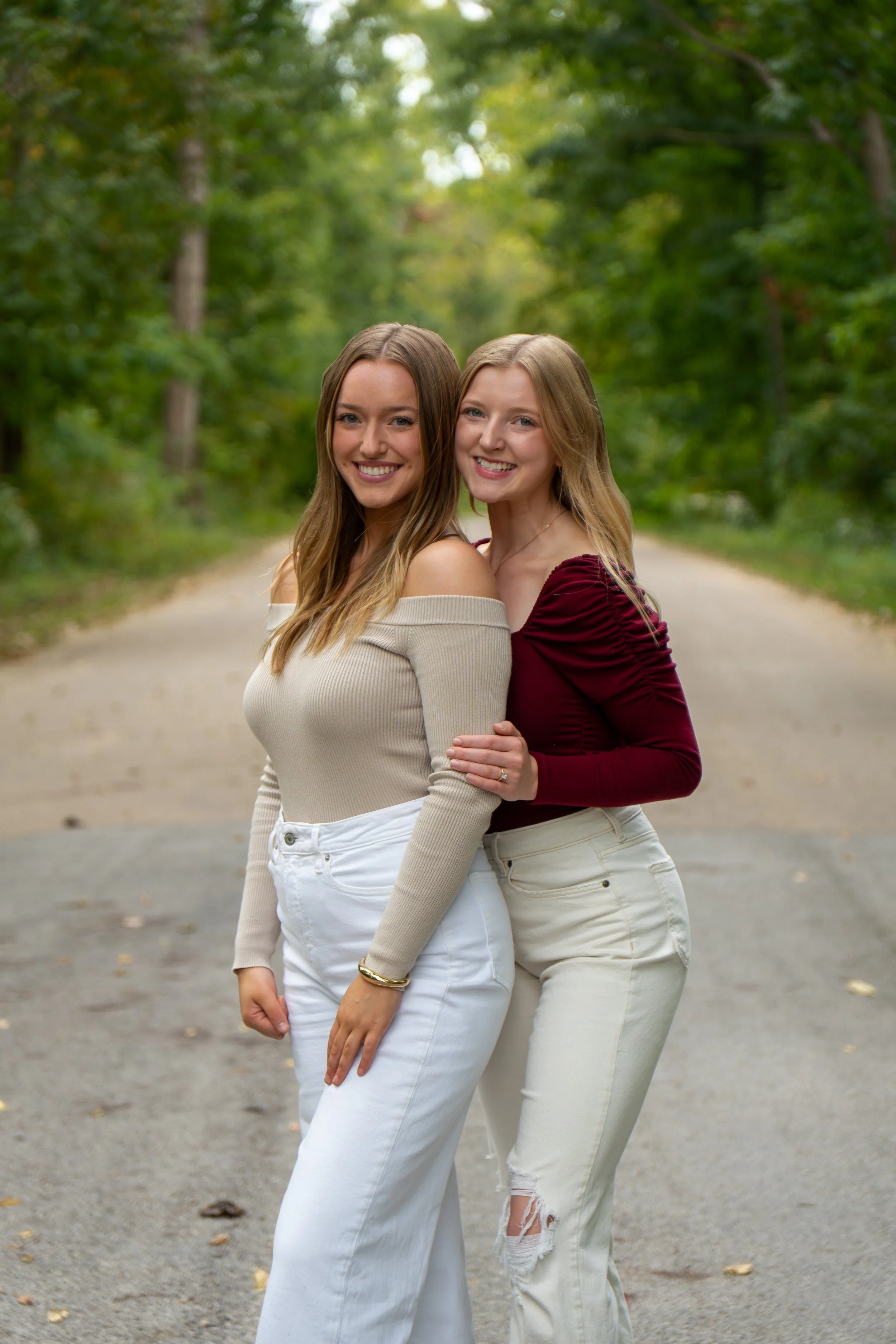 Two young women standing close together on a tree-lined path, smiling at the camera. They are wearing casual outfits, with one in an off-shoulder beige top and white jeans, and the other in a maroon long-sleeve top and distressed white jeans. The bac