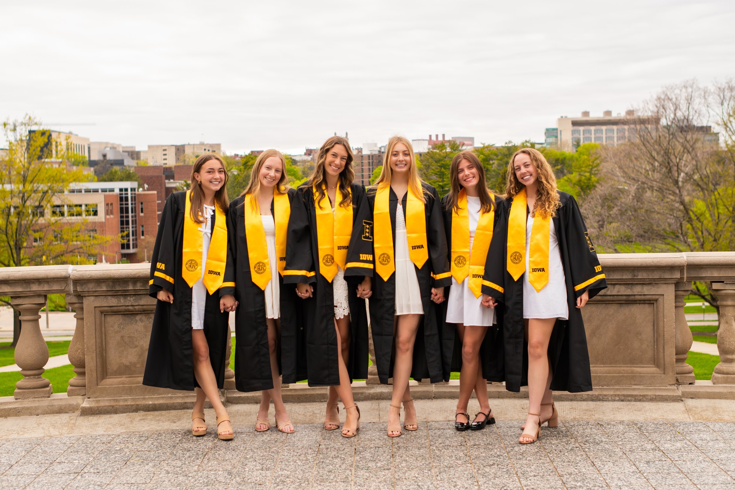 Six women in graduation gowns and yellow stoles standing together outdoors on a stone balcony with a cityscape and overcast sky in the background.