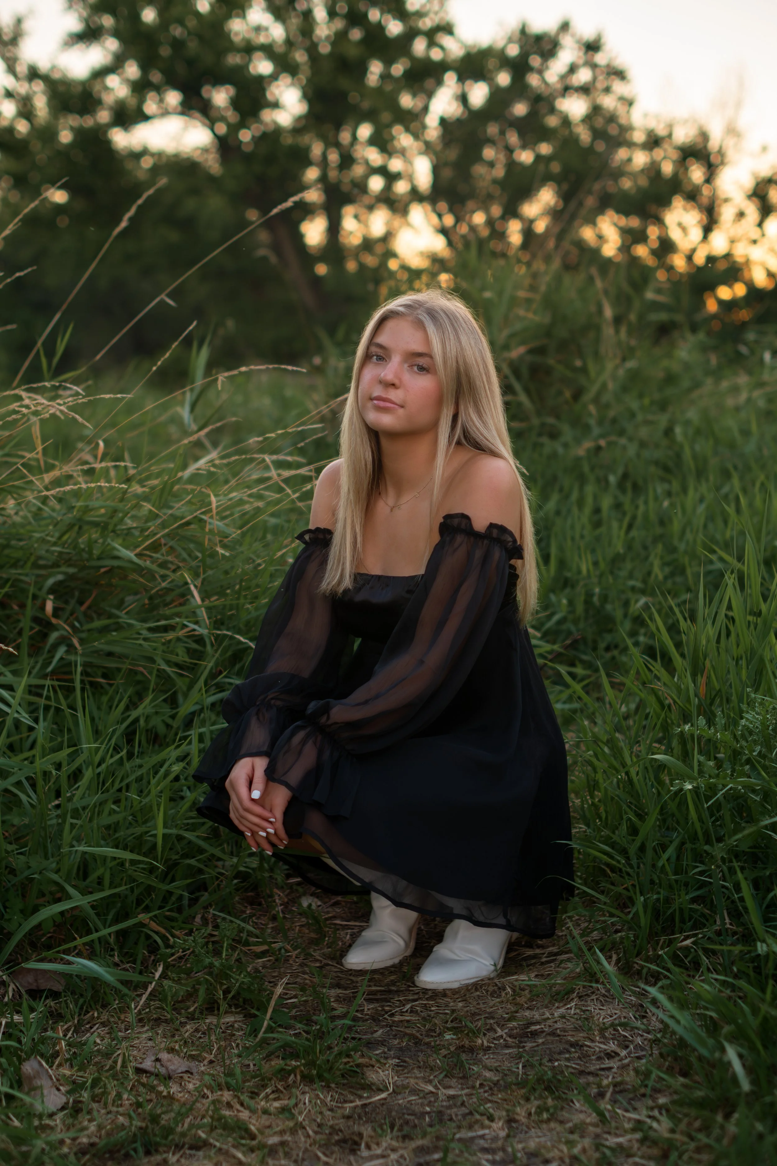 A young woman with long blonde hair, wearing a black dress with sheer sleeves, sitting in a grassy outdoor setting during sunset.