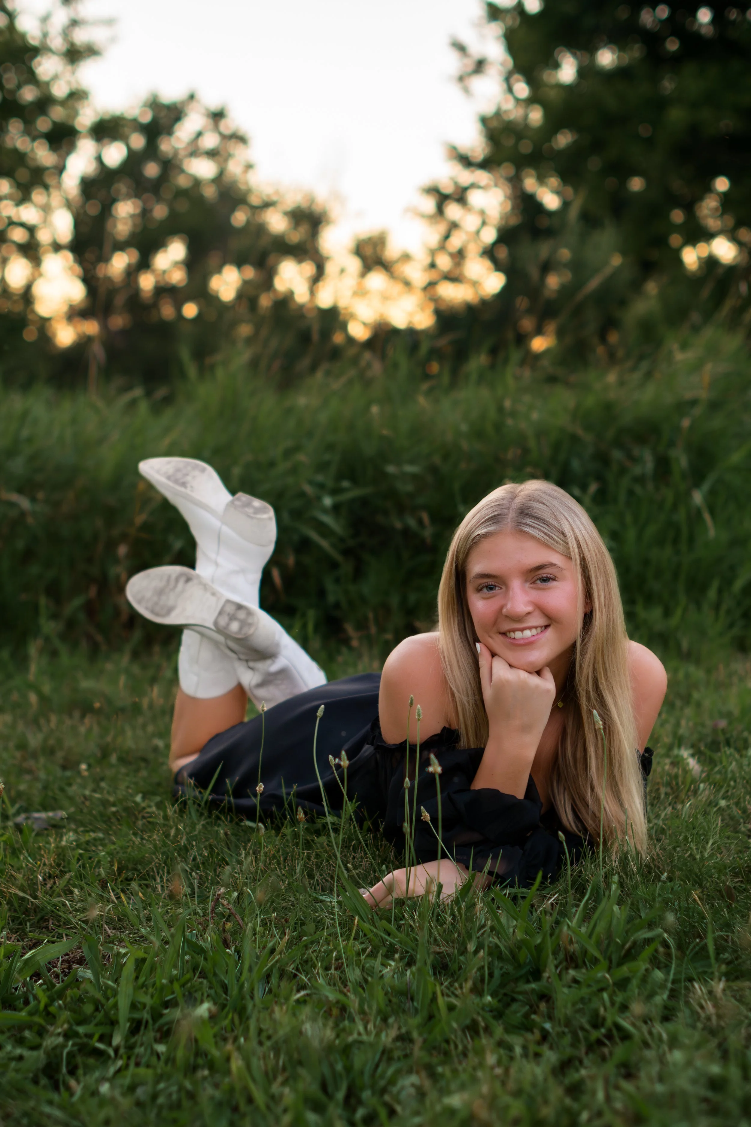 A young girl with long blonde hair lying on her stomach in grass, smiling, with a blurred green trees and sky in the background.