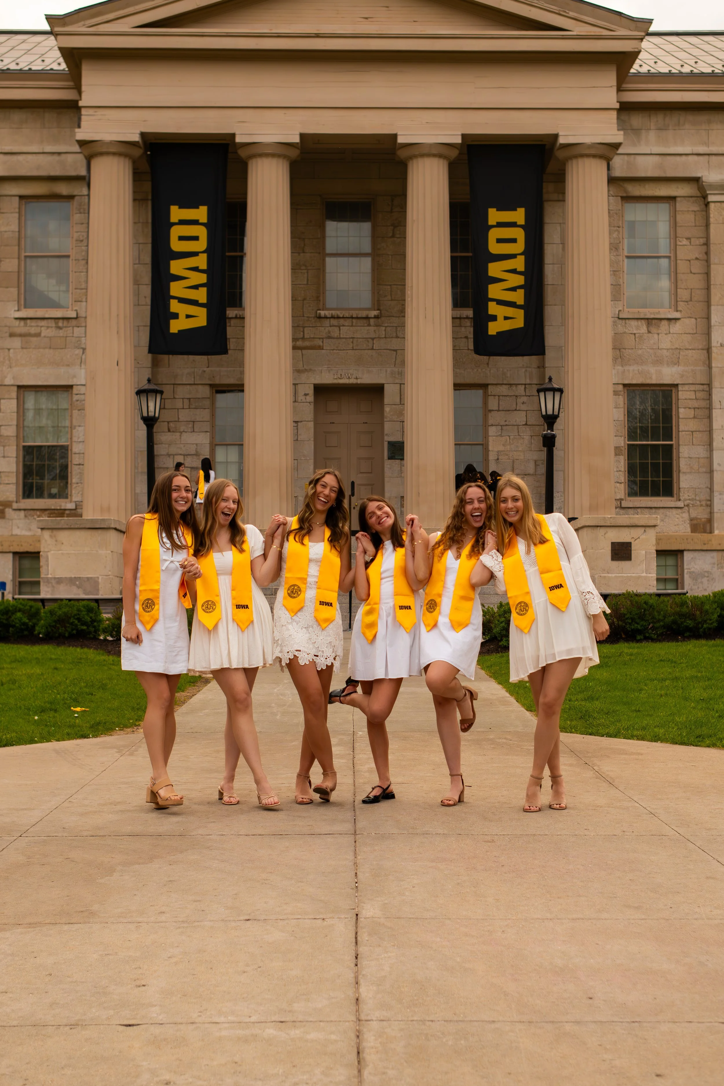 A group of seven young women wearing white dresses and yellow academic stoles pose happily in front of a university building with large columns and black banners that say "IOWA" in yellow.