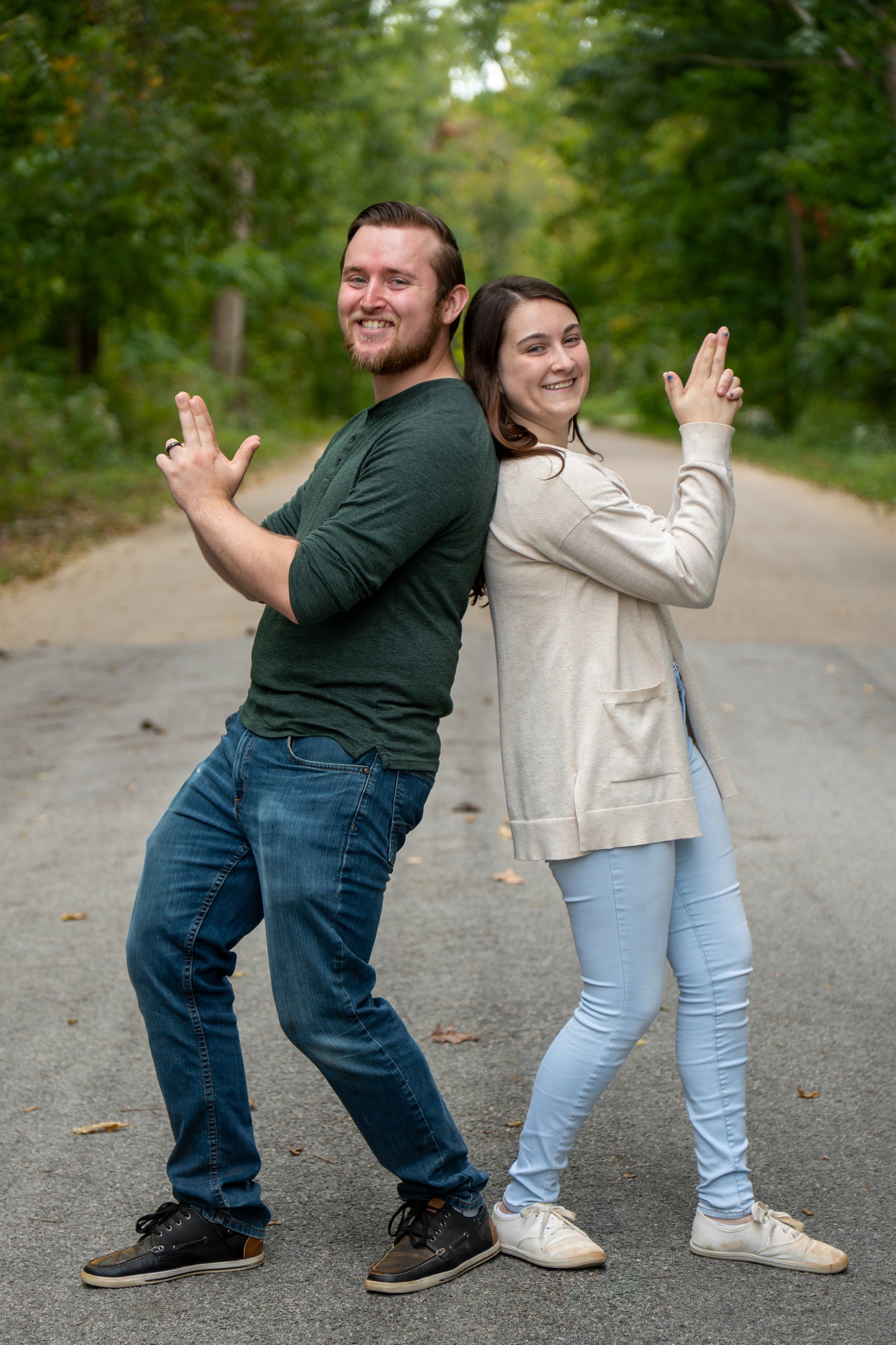 Two young adults, a man and a woman, stand back-to-back on a forest path, each smiling and pointing a finger gun in opposite directions.