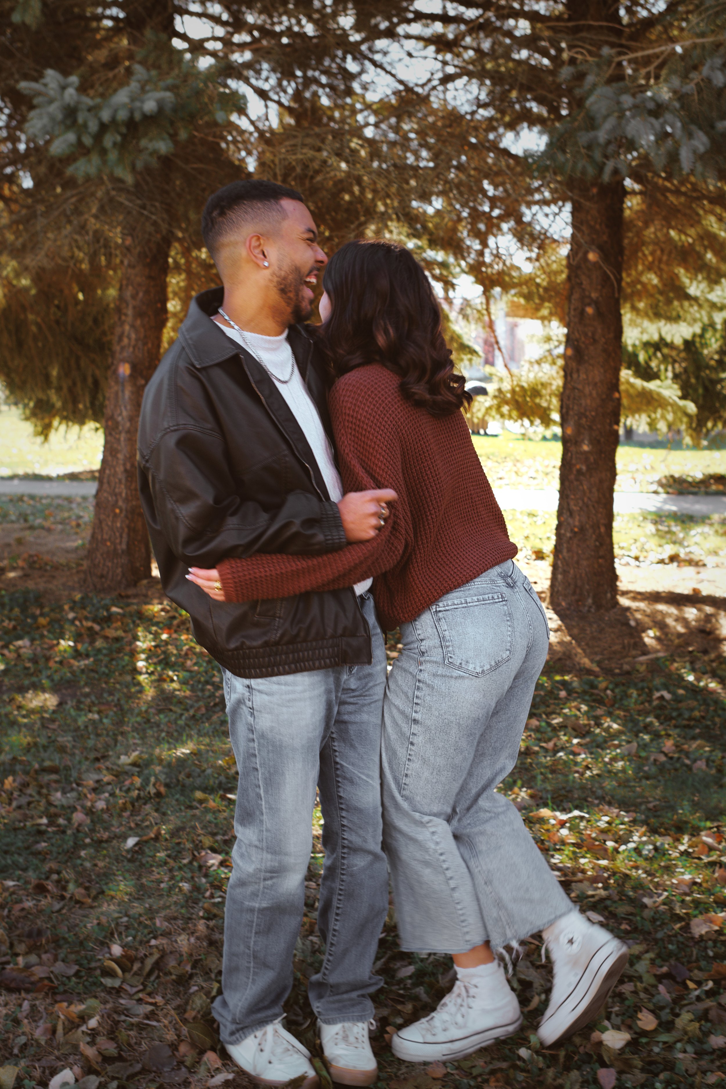 Two people, a man and a woman, are standing close together outdoors under a large evergreen tree. They are smiling and appear to be happy. The man is wearing a black jacket, white t-shirt, and light-colored jeans. The woman is wearing a red sweater, 