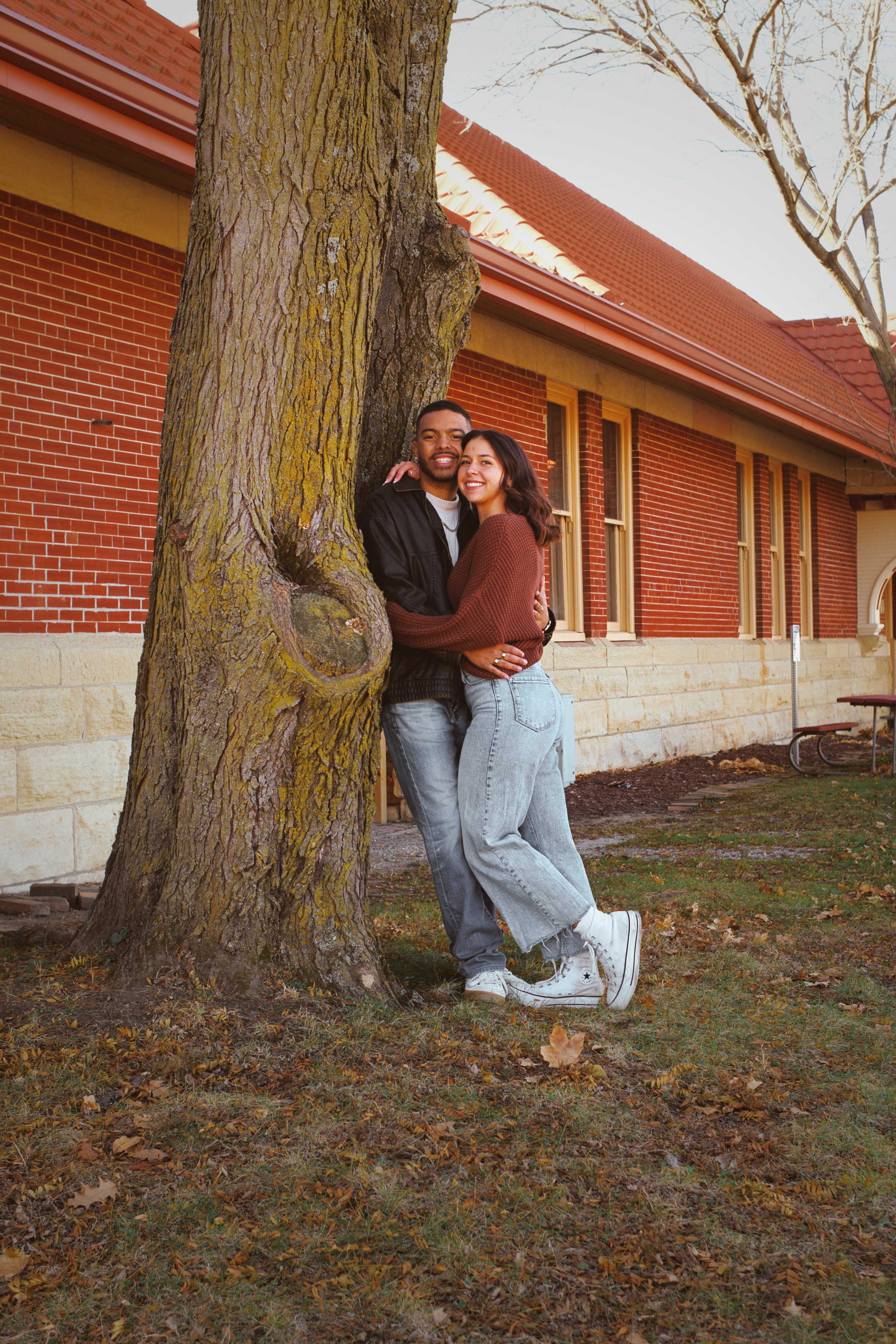 A young couple hugging in front of a large tree on a fall day, with a brick building and leafless trees in the background.