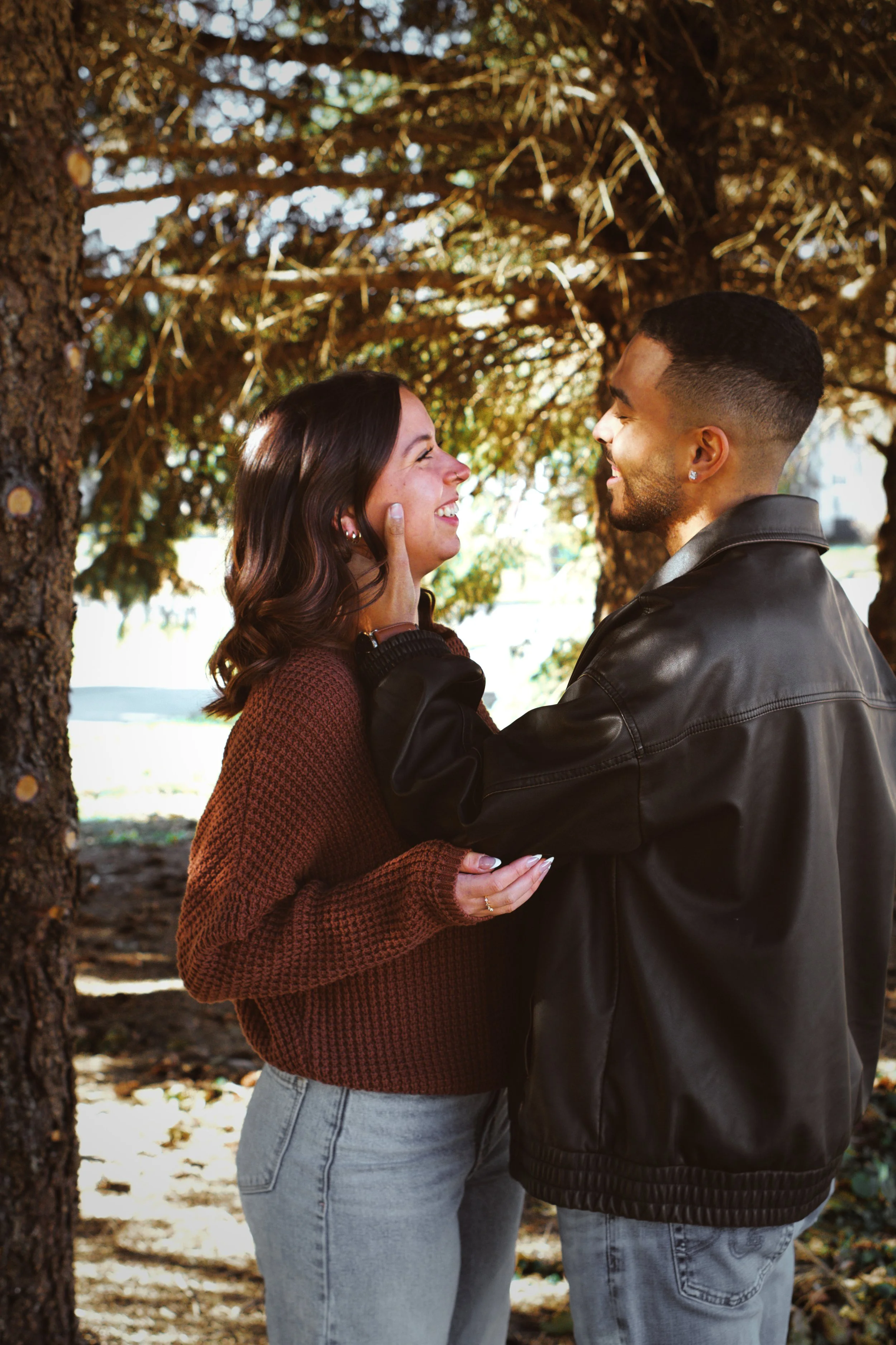 A couple standing closely together outdoors under a large tree, smiling at each other, with the woman's hand touching the man's face, both appearing happy.