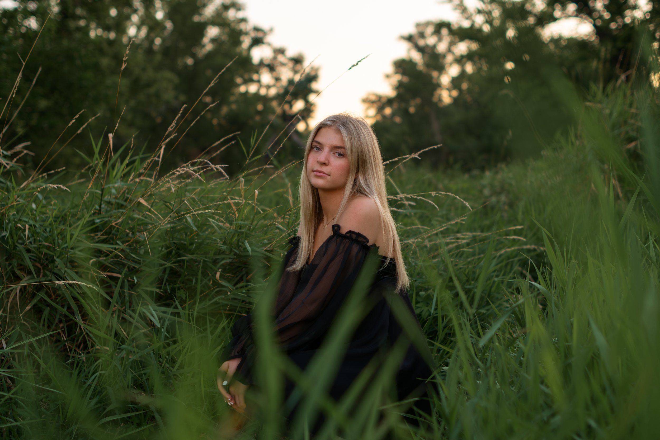 Young woman with long blonde hair sitting in tall green grass with trees and a bright sky in the background.