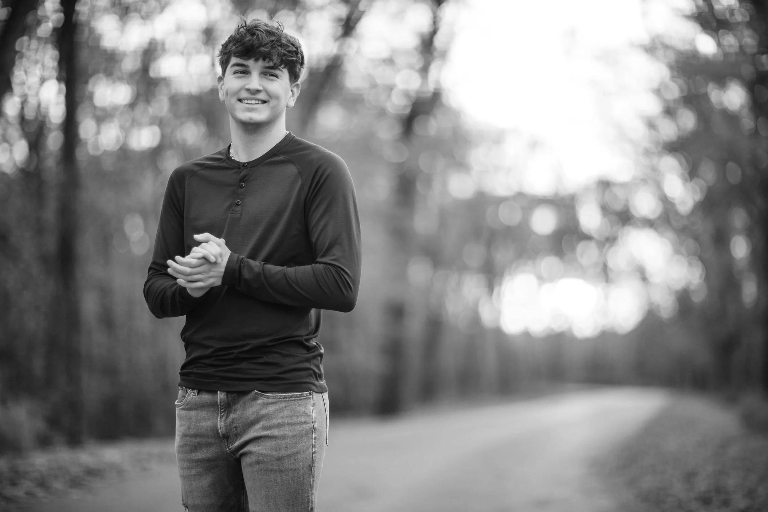 A young man smiling outdoors in a park, wearing a long-sleeve shirt and jeans, with trees and blurred sunlight in the background.