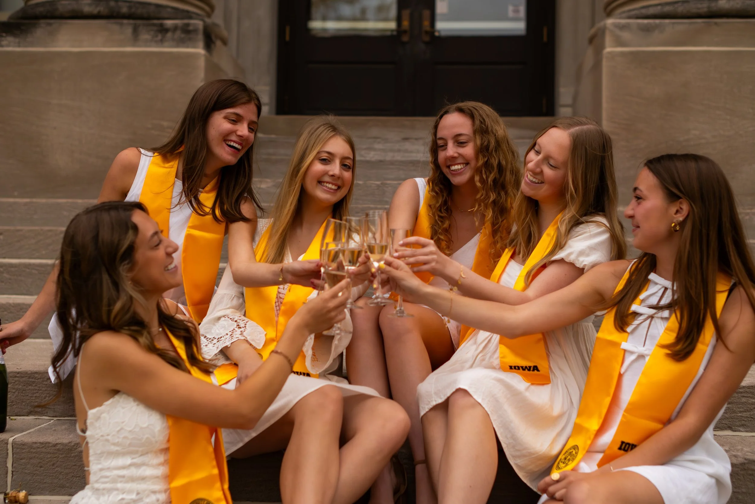 Group of young women wearing white dresses and yellow sashes sitting on steps, laughing and toasting with champagne glasses.