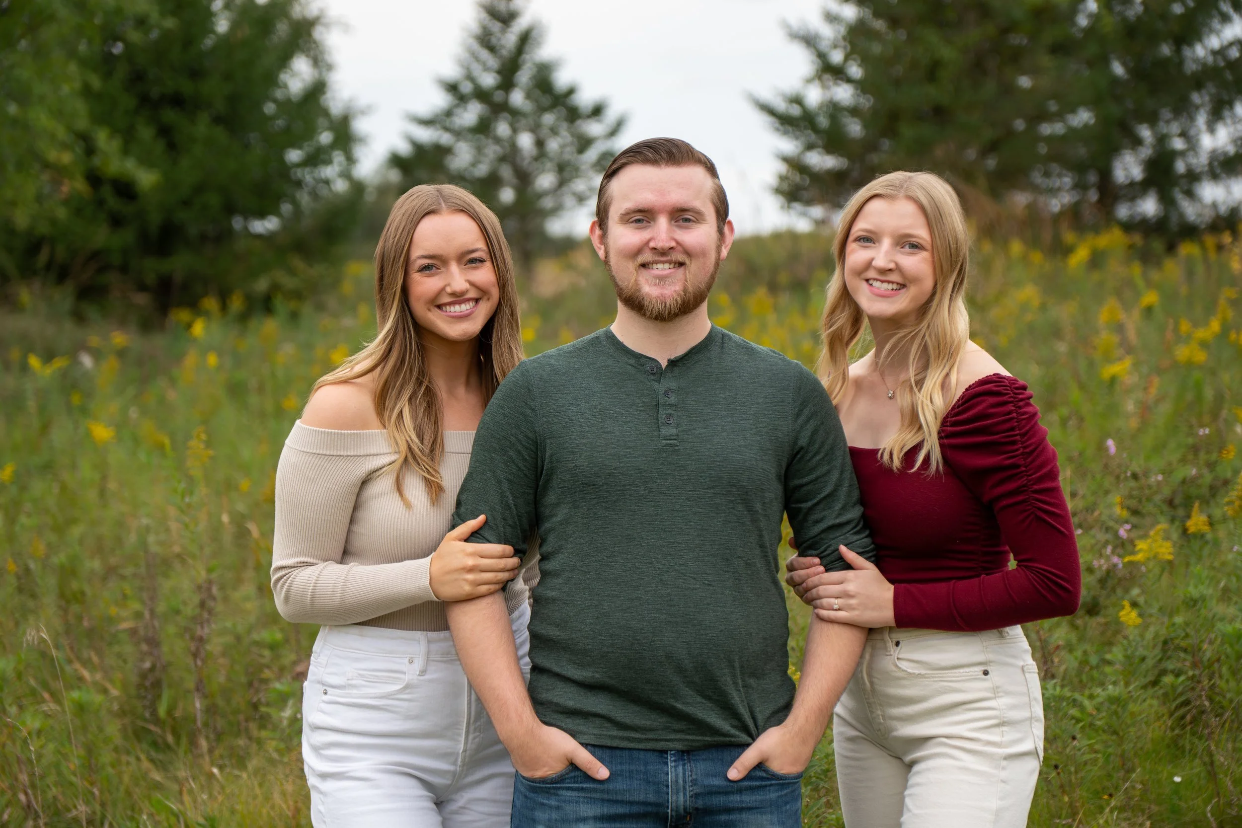 Three young adults, two women and one man, smiling and standing outdoors in a field with trees and yellow flowers in the background.
