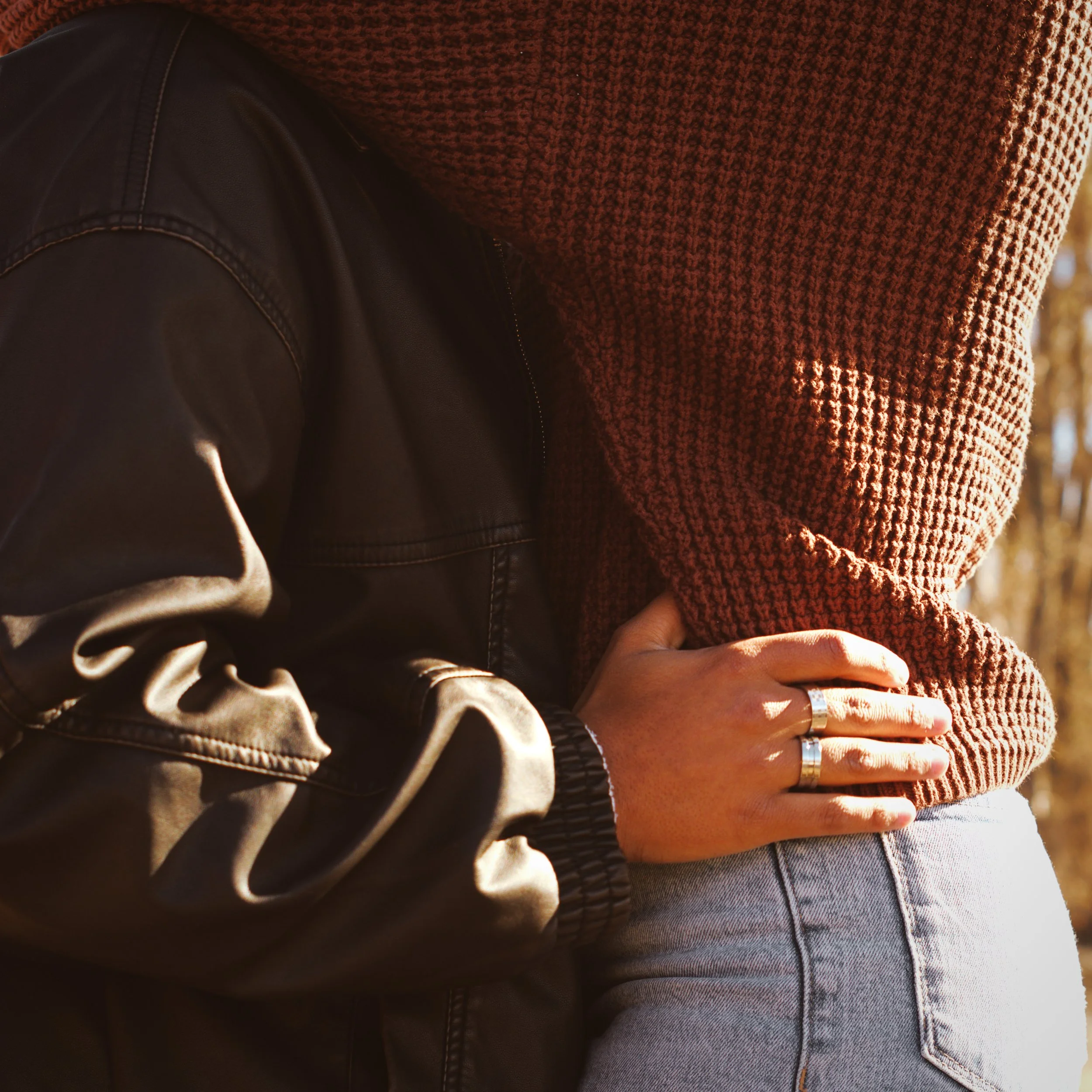 A close-up of a person wearing a brown knit sweater, gray pants, and a silver ring on their finger, with a leather jacket partially visible nearby.