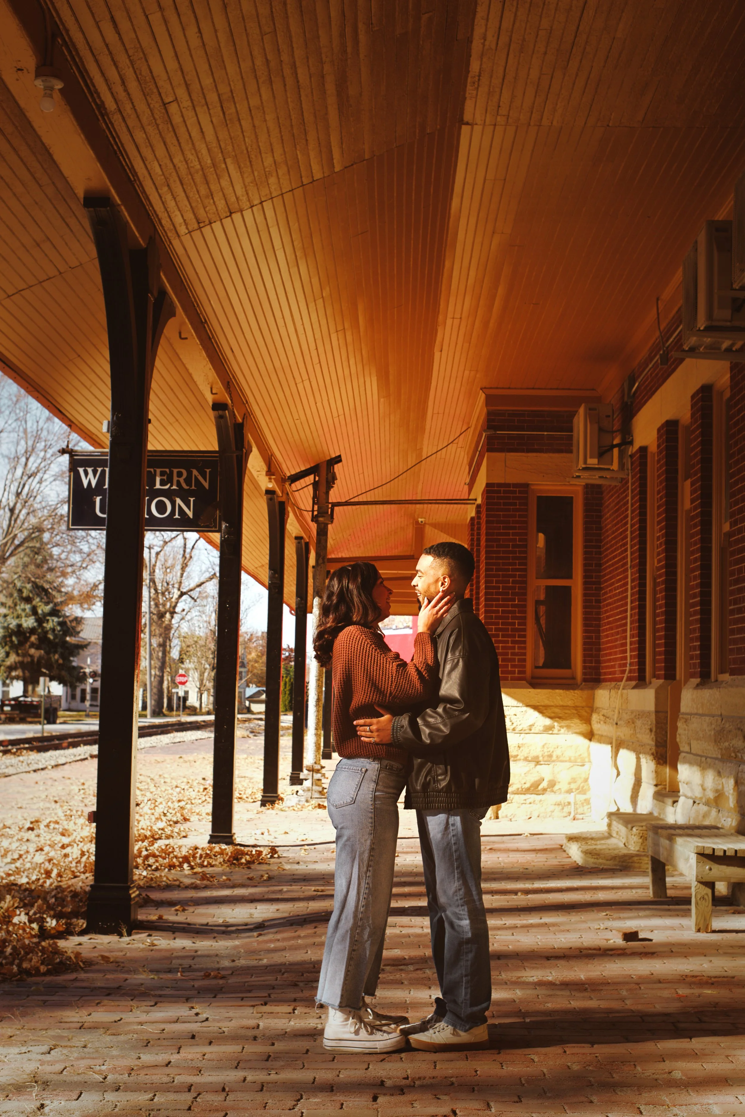A couple stands on a brick sidewalk, facing each other and holding hands. They are under a wooden awning outside a historic brick building. The scene is during autumn, with fallen leaves on the ground and trees with sparse leaves in the background.