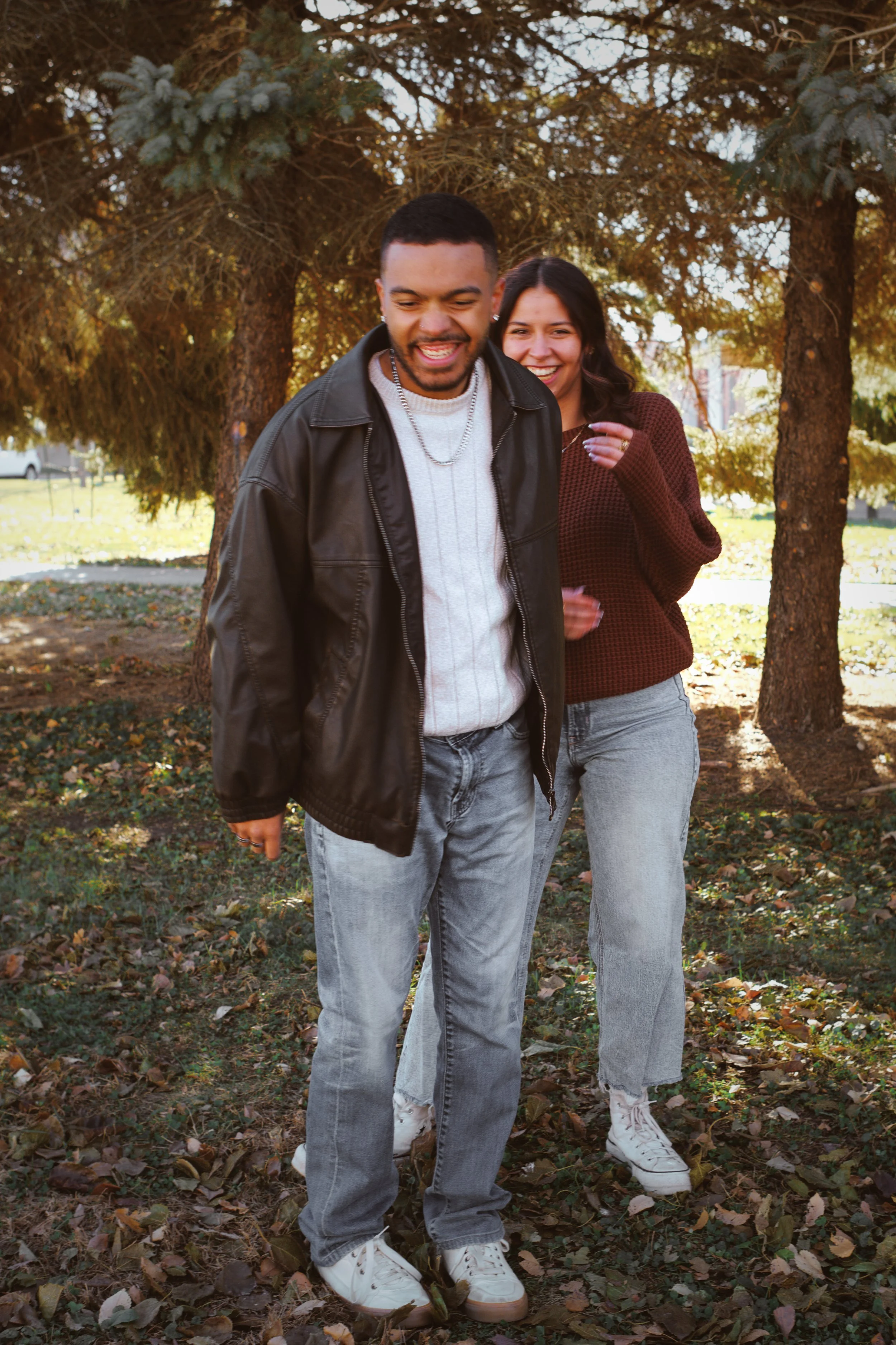 A man and a woman standing outdoors on a fall day, smiling and laughing. The man is wearing a black leather jacket, white sweater, and jeans. The woman is wearing a brown sweater and light jeans. They are surrounded by trees with golden leaves, and t