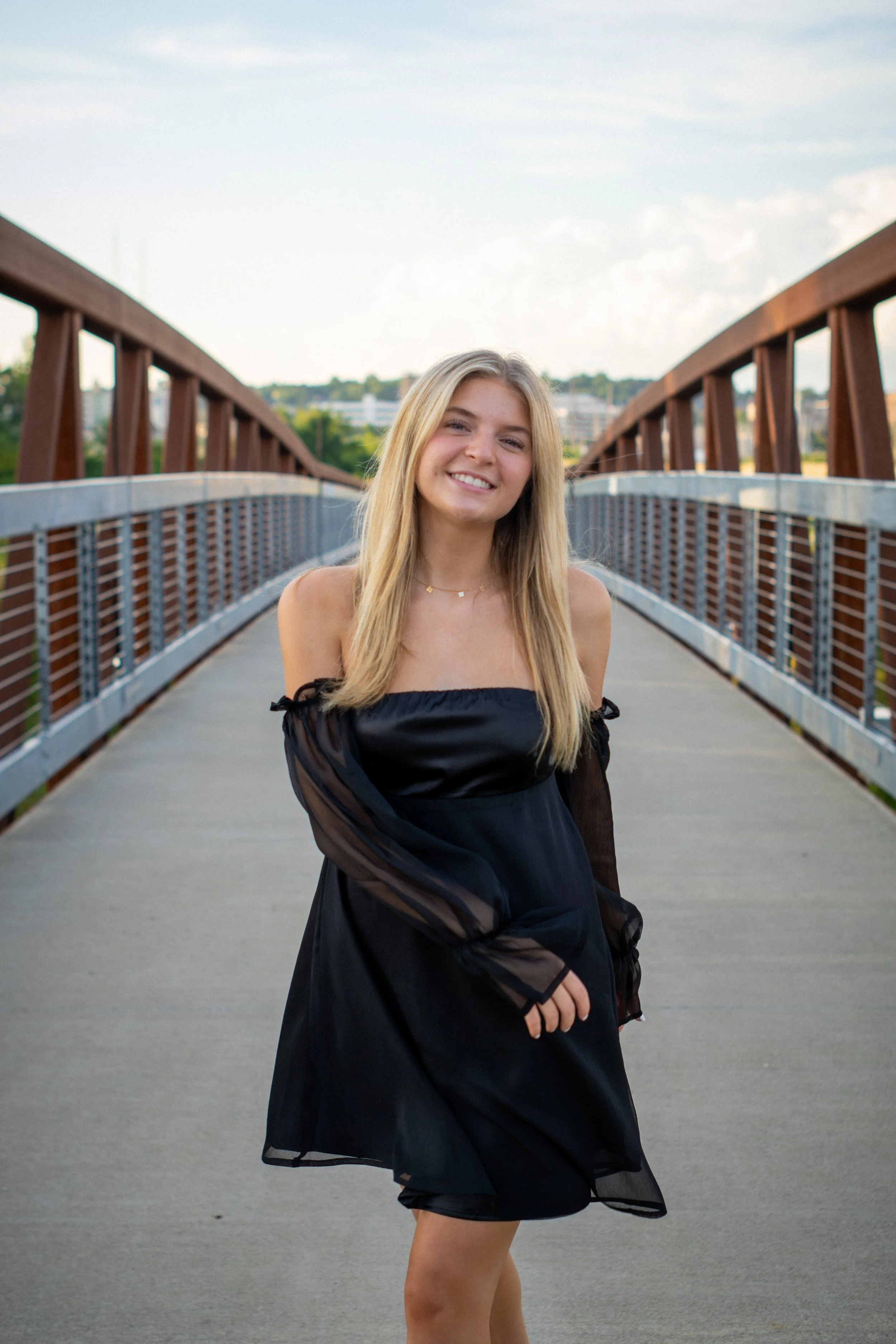 A young woman with blonde hair, smiling, stands on a bridge with a wooden railing, wearing a black dress with sheer sleeves, outdoors during daytime.