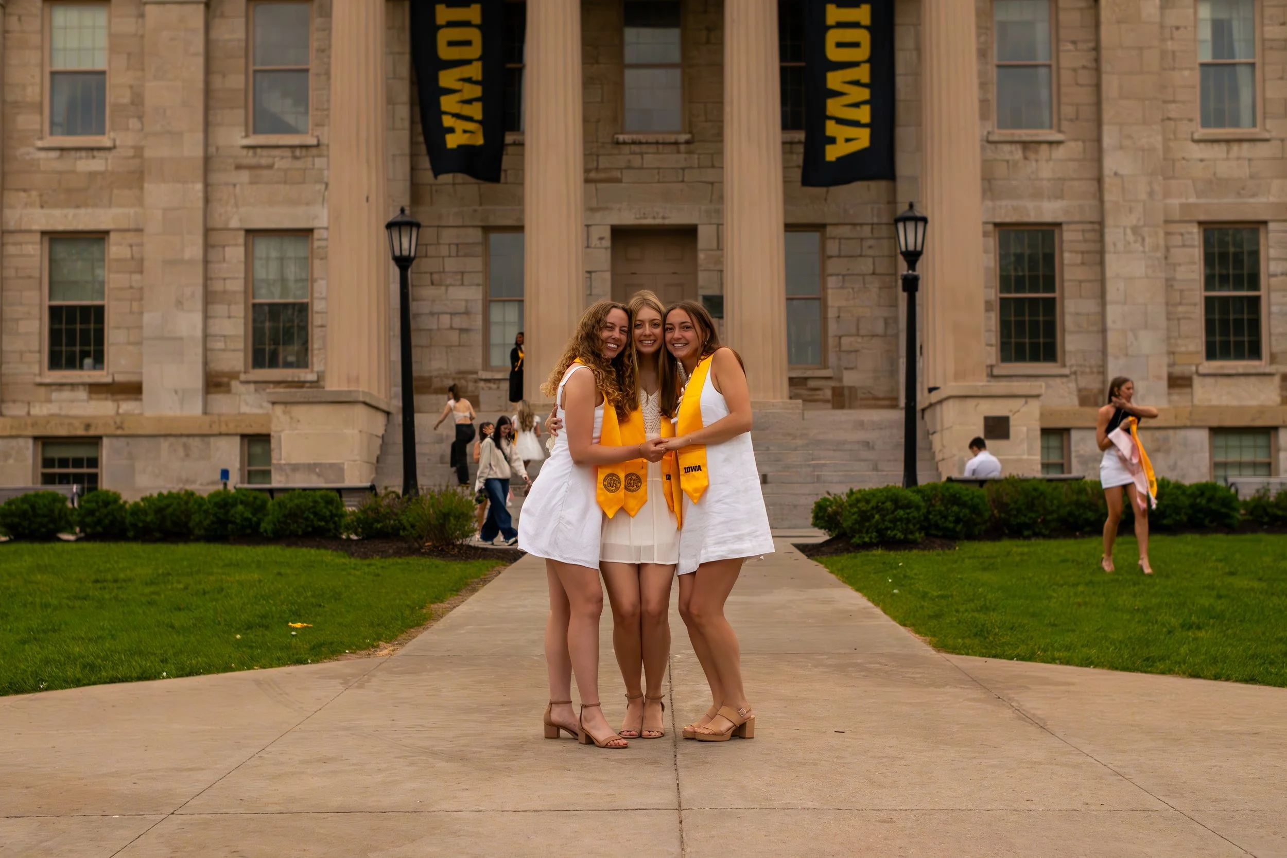 Three young women are standing close together on a walkway in front of a university building, smiling at the camera. They are wearing white dresses and yellow graduation stoles, indicating a graduation celebration.