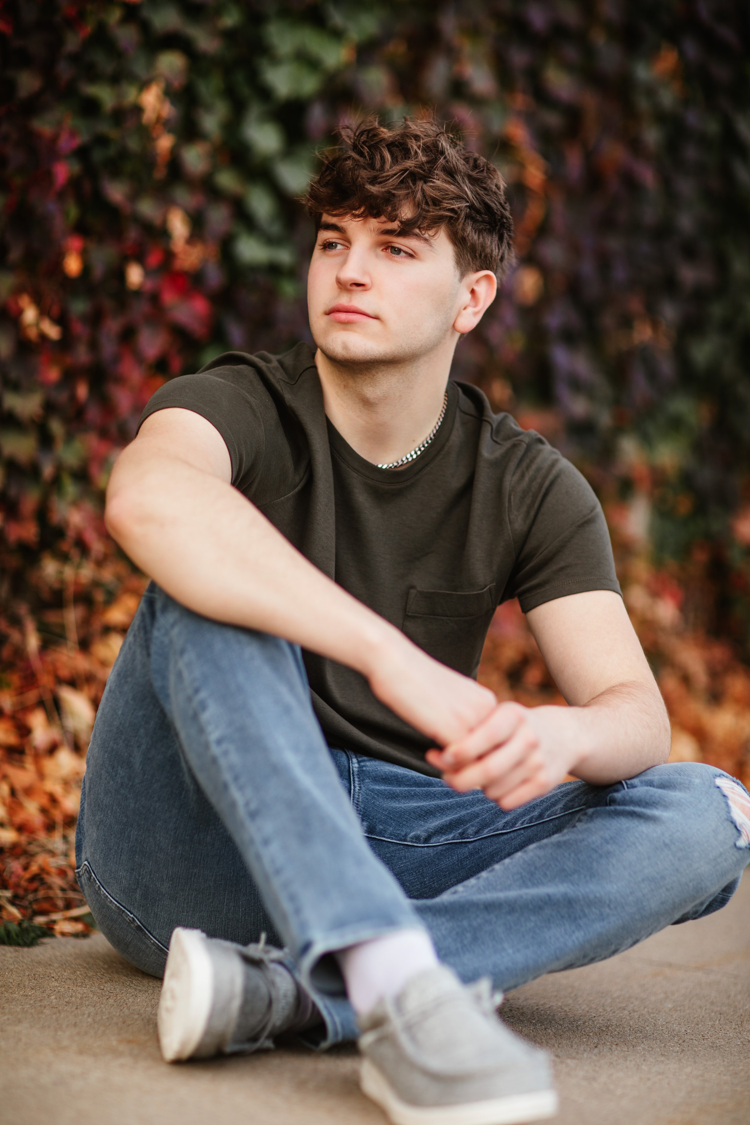 A young man with curly brown hair wearing a black t-shirt and blue jeans, sitting outdoors on ground with fallen leaves, looking to the side.