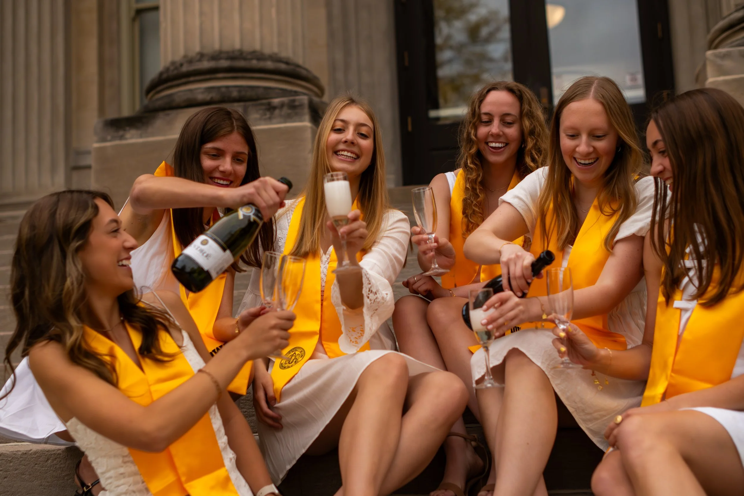 A group of young women sitting on steps, smiling, and celebrating with champagne, wearing yellow sashes and white outfits outside a building with stone columns.