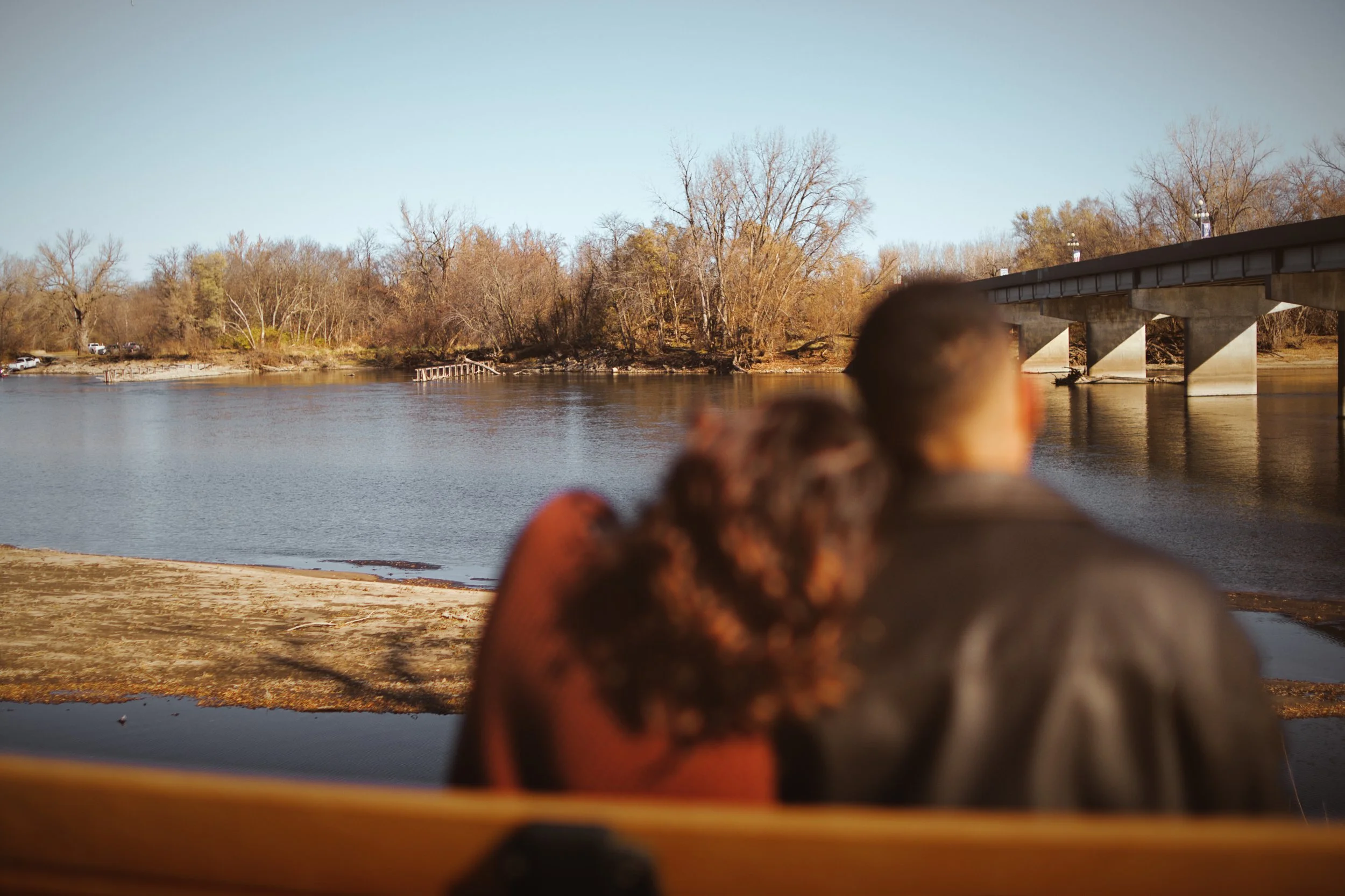 A person standing on a sandy beach near a body of water, with a bridge overhead and trees on the opposite bank.
