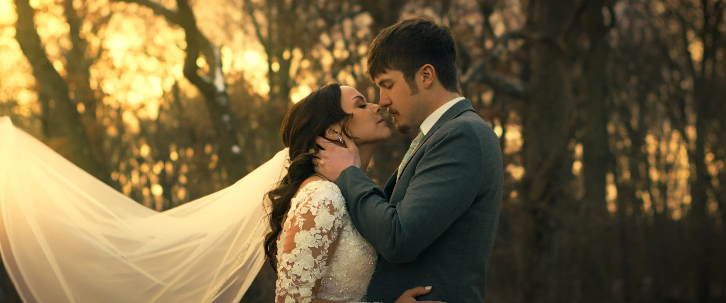 A bride and groom embrace outdoors during sunset, with the bride wearing a lace wedding dress and veil, and the groom in a suit, as they look into each other's eyes.