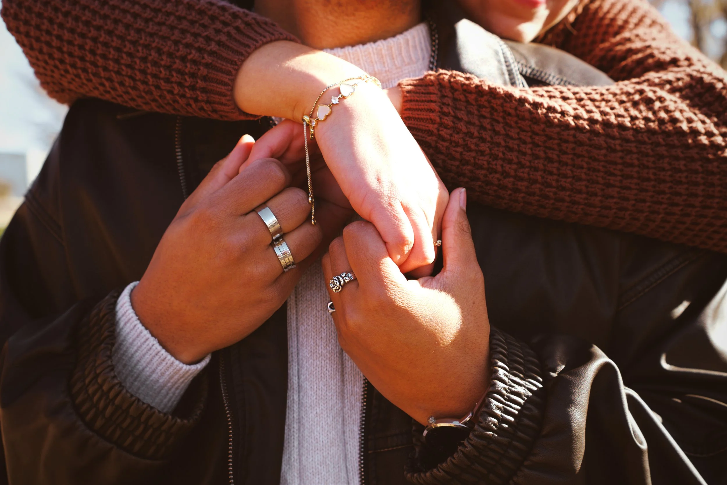 Two people holding hands, one wearing a brown knit sweater and multiple rings, the other wearing a black jacket with metal zipper and jewelry. The photo focuses on their hands and bracelets.