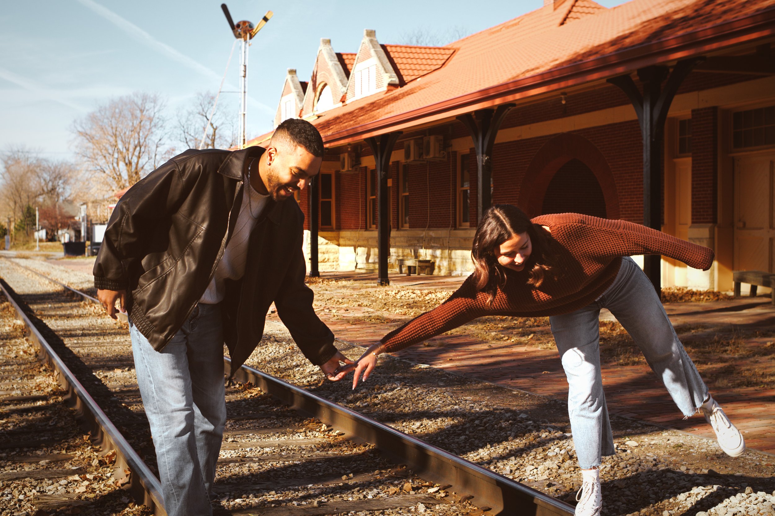 Two people, a man and a woman, are leaning over and working on a railroad track outdoors on a sunny day. There are buildings with red roofs and trees in the background.
