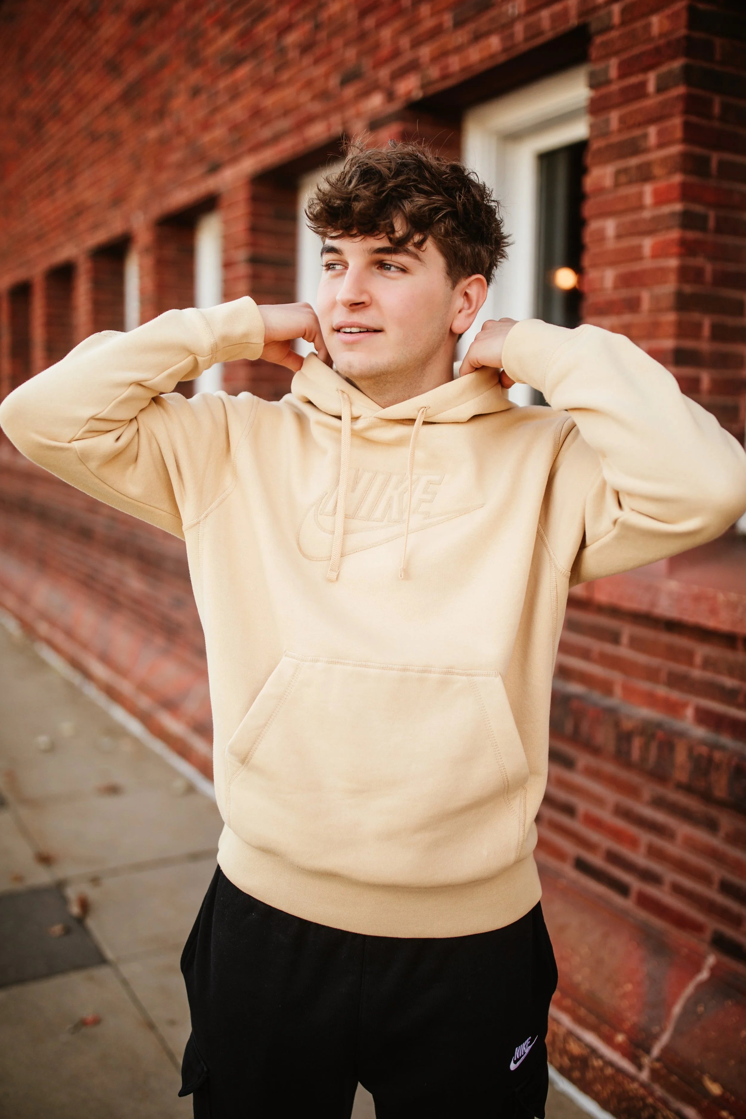 Young man in a beige hoodie and black Nike sweatpants adjusting his hoodie outside next to a brick building.