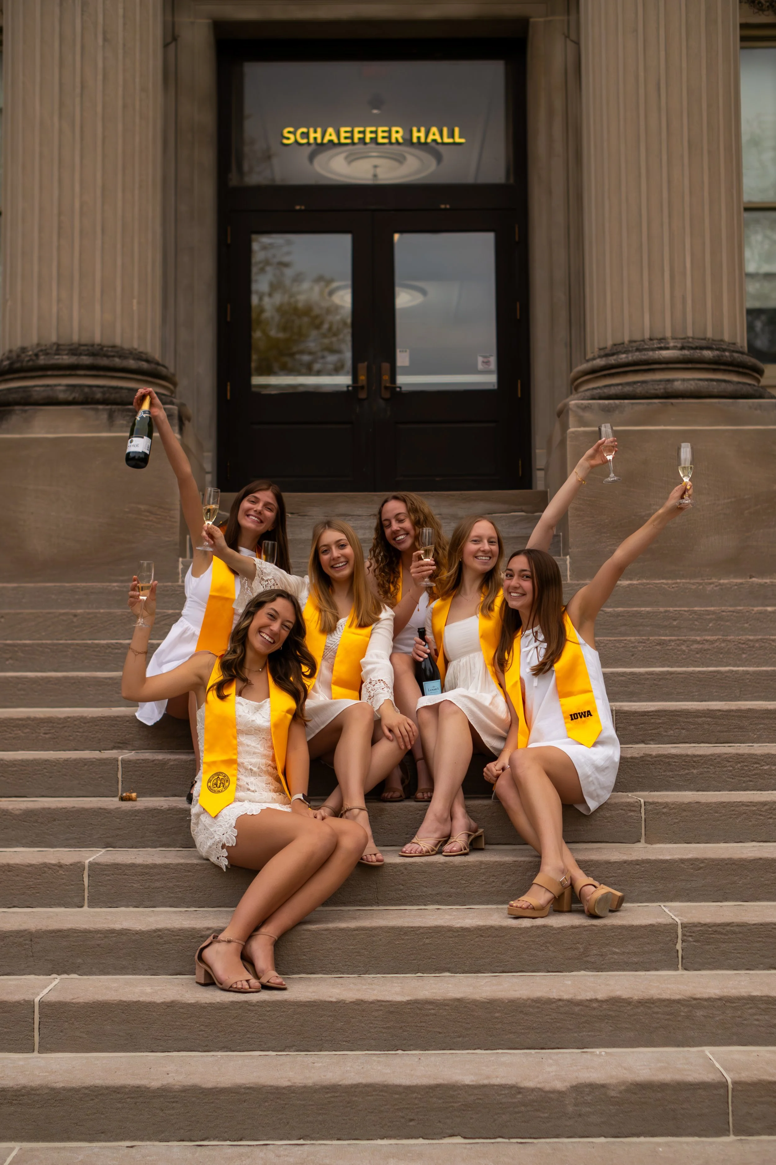 A group of young women celebrating graduation on the steps of a building labeled 'Schaeffer Hall.' They are dressed in white clothing with yellow sashes and are holding champagne glasses and bottles, smiling and toasting.