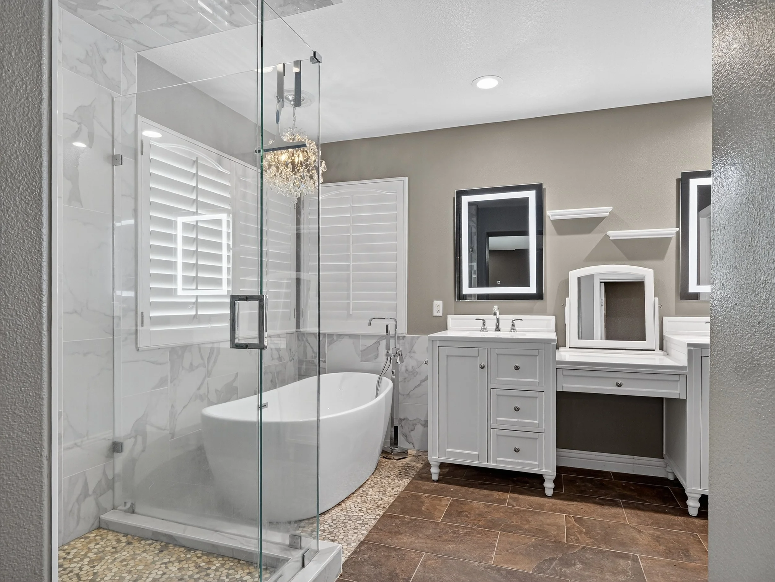 A modern bathroom featuring a glass-enclosed shower with a white freestanding bathtub inside. The bathroom has a vanity with a white cabinet, a rectangular mirror with a light frame, and floating shelves. The wall behind the vanity is painted a light