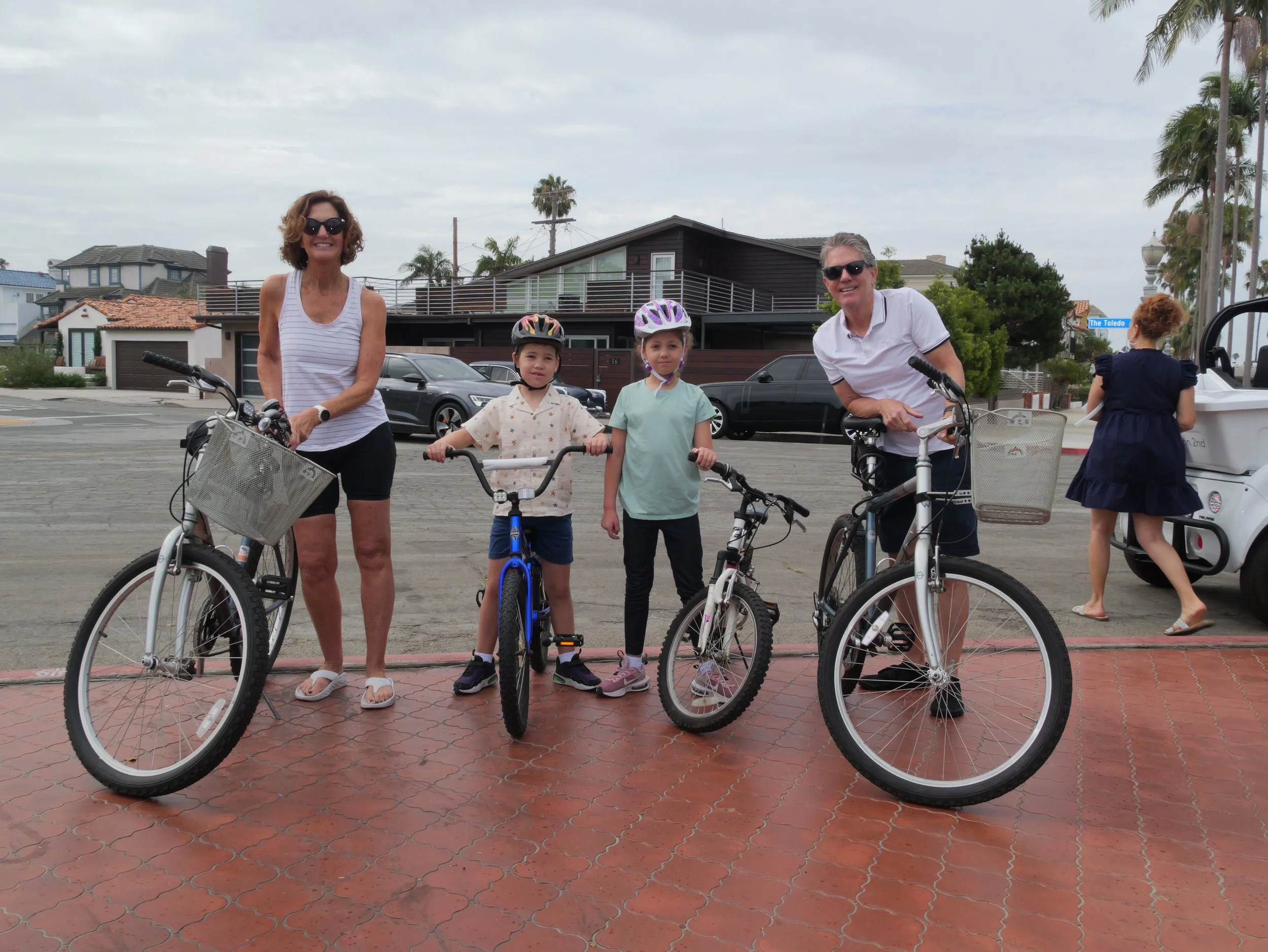 A group of six people, including two adults and four children, stand with bicycles on a city street, smiling at the camera. The background features houses, parked cars, palm trees, and a street sign reading "The Toledo." The sky is overcast.