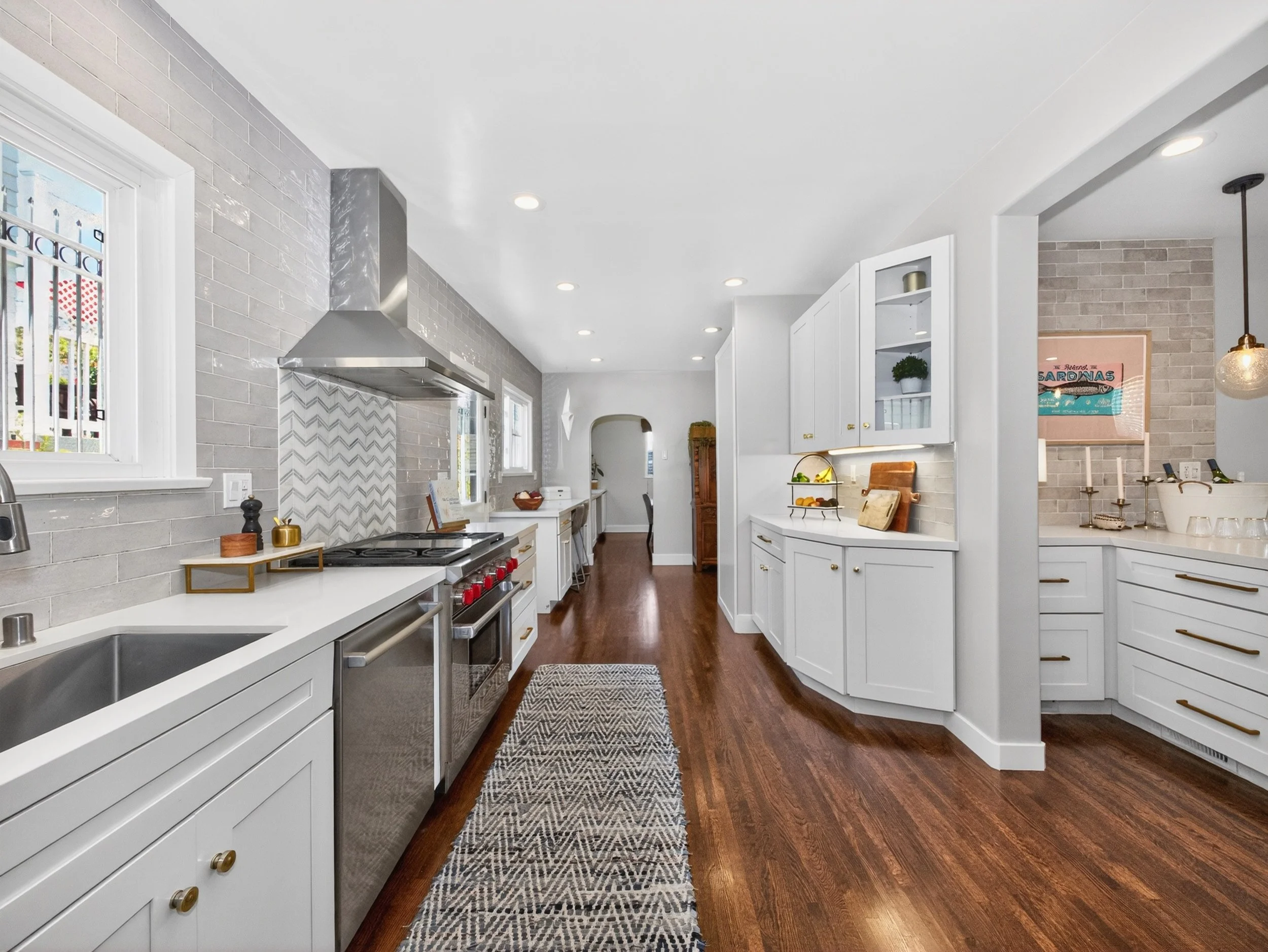 A bright, modern kitchen with white cabinets, gray subway tile backsplash, stainless steel appliances, and hardwood flooring. Natural light streams through the windows, illuminating the space.