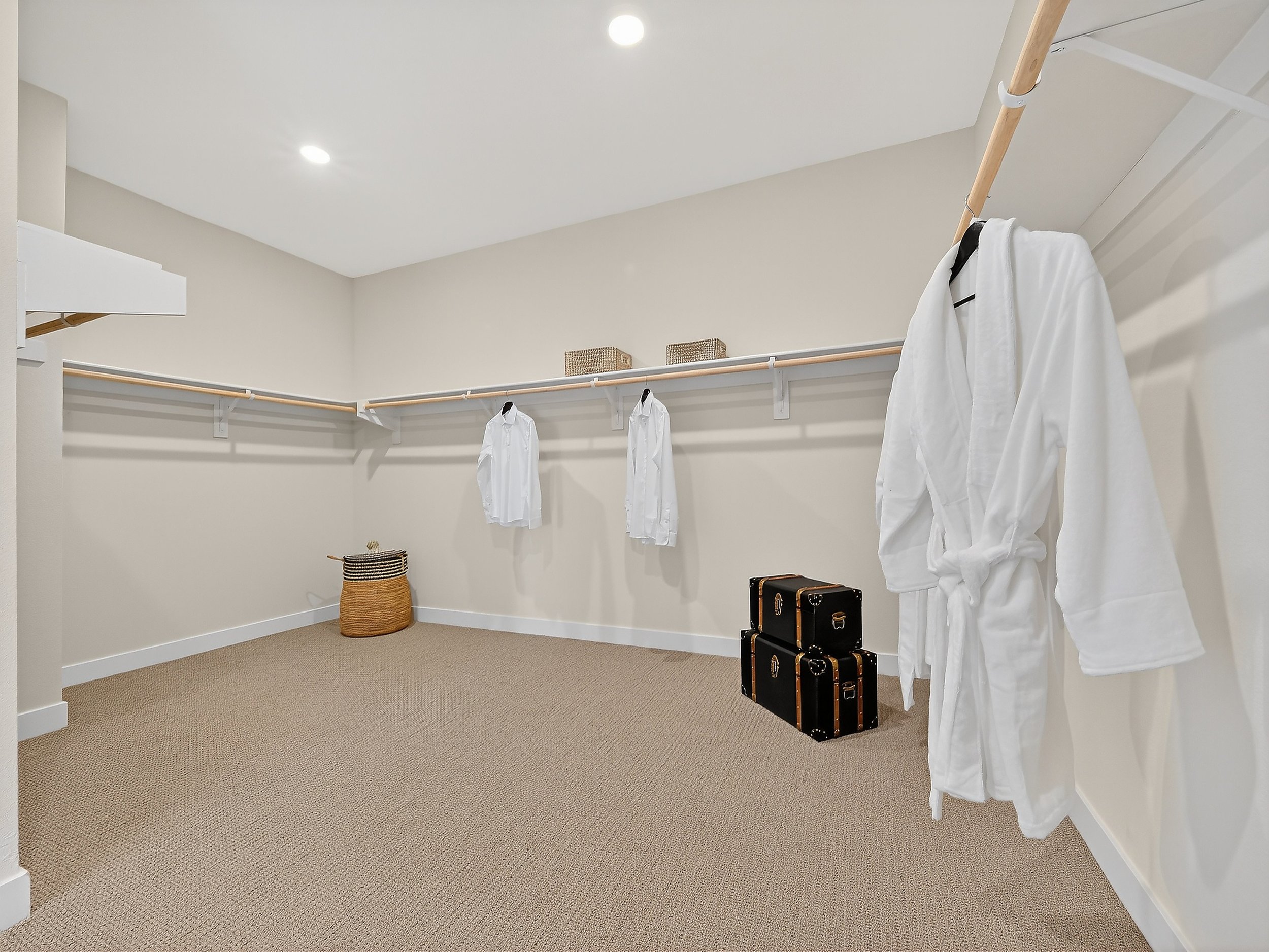 A walk-in closet with white walls and carpeted floor, featuring a hanging rod with white shirts, a wicker basket on the floor, and two hard-shell suitcases.