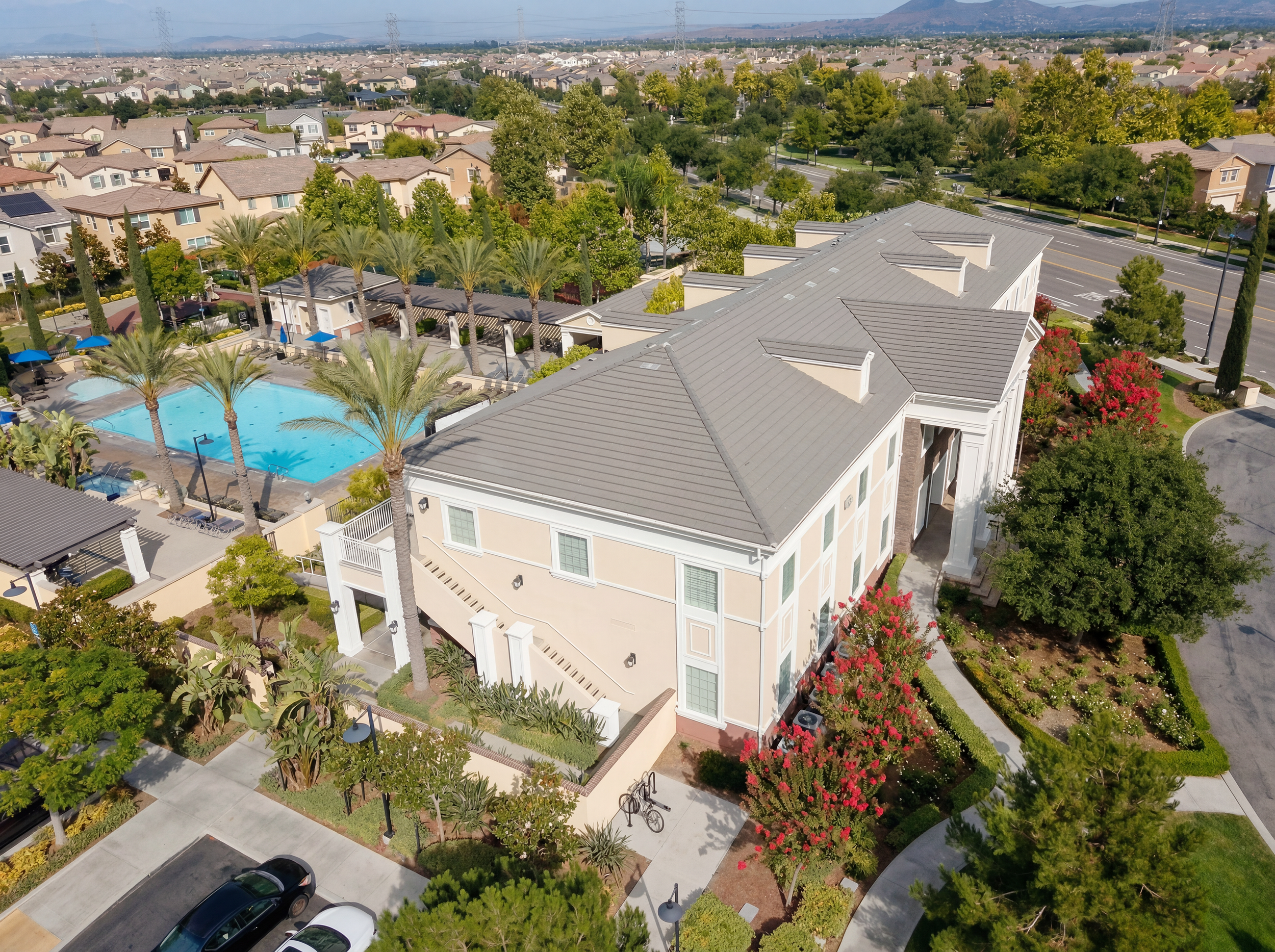 Aerial view of a residential area with a swimming pool, palm trees, and modern houses, surrounded by greenery and parking lots.