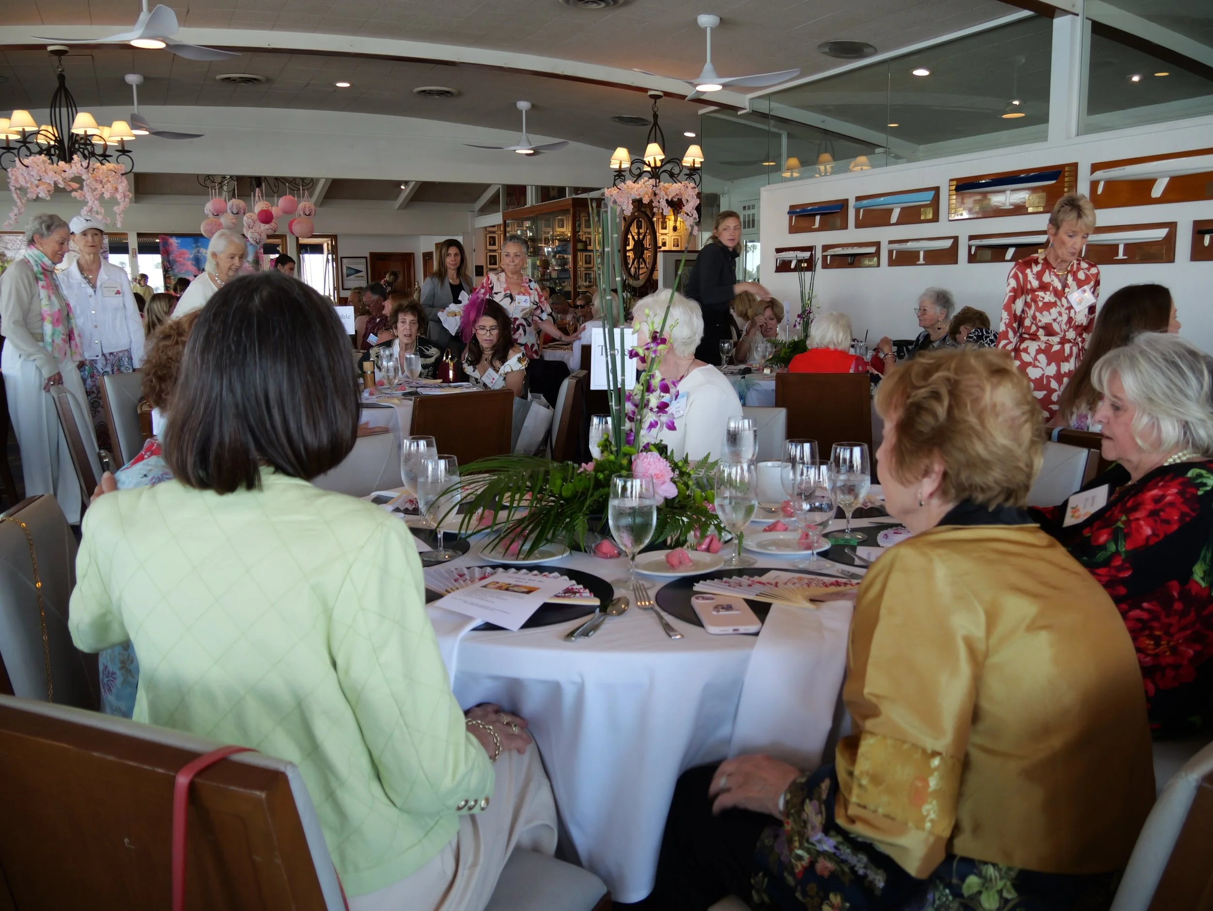 A group of elderly women gathered around a dining table at a social event inside a restaurant. The table is decorated with flowers and surrounded by women wearing colorful and patterned clothing. There are pink paper lanterns and flowers hanging from