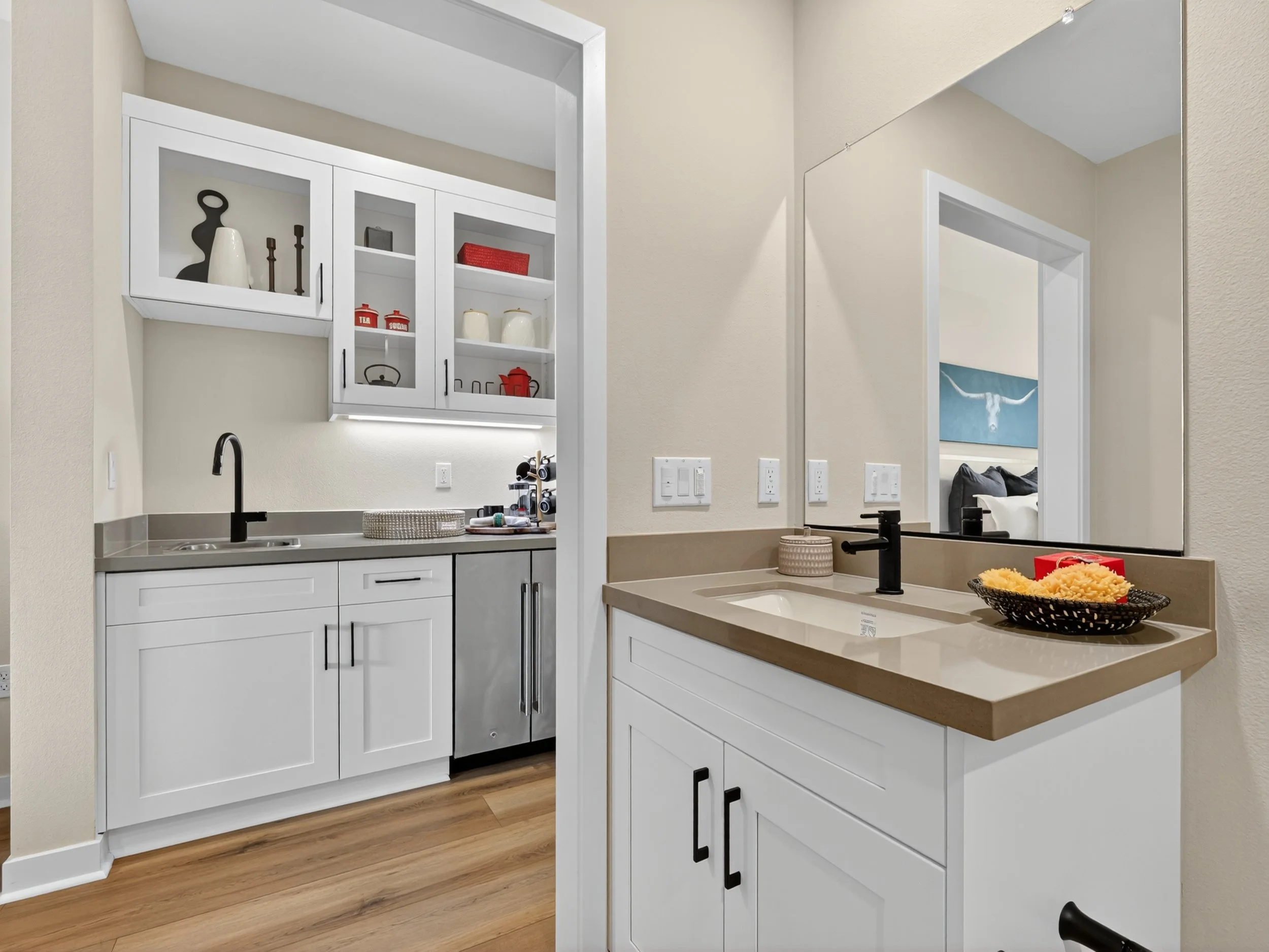 A modern kitchen with white cabinets, a beige countertop, and a large mirror on the wall. The backsplash has a small window and some decorative items, including a black sculpture and candlesticks. There are electrical switches on the wall, and some kitchen items are placed on the counter.
