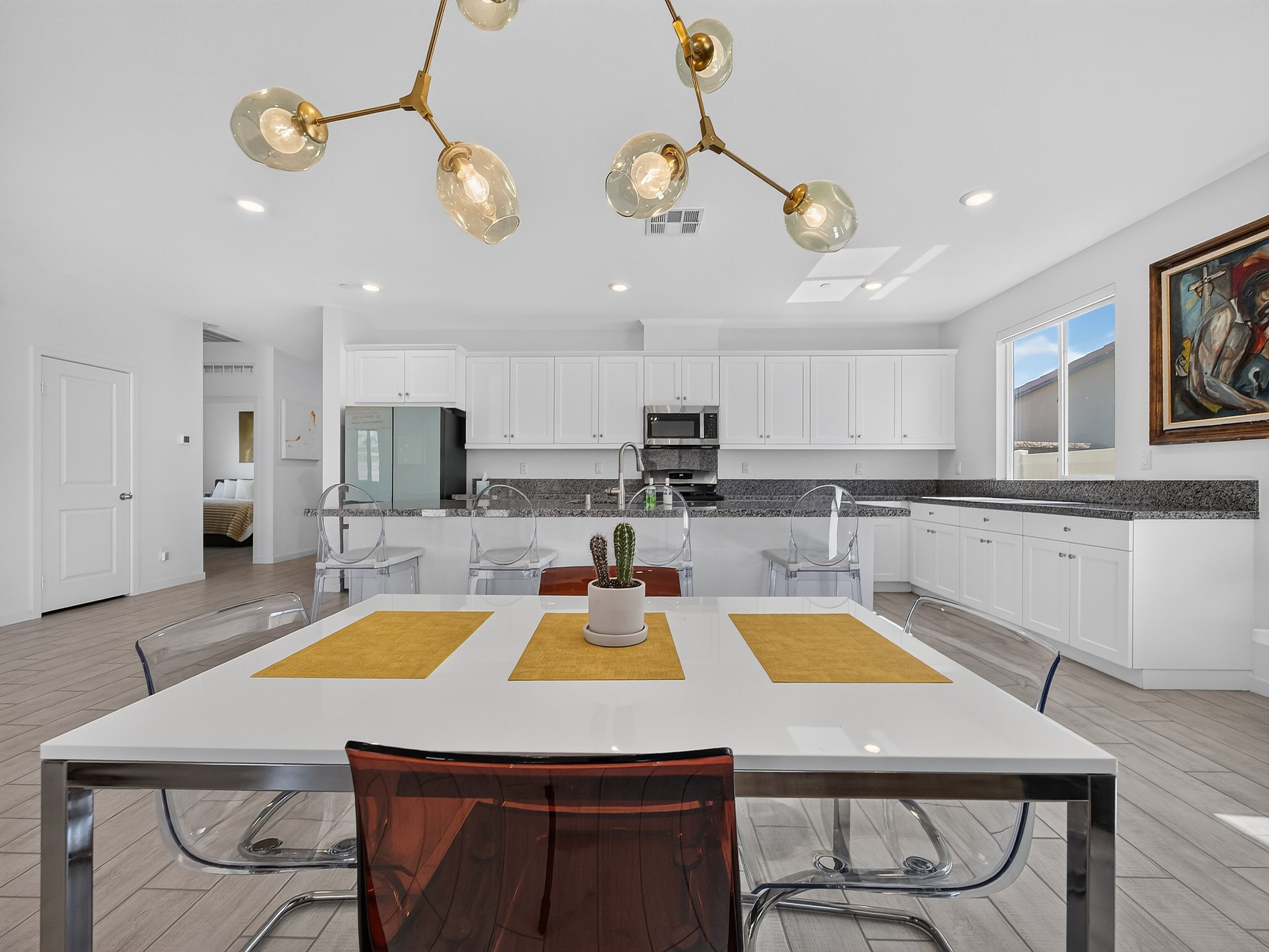 A modern kitchen with white cabinets, gray countertops, a microwave above an oven, and a large window with blinds. A dining table with a white top and mustard placemats is in the foreground, surrounded by transparent and dark amber chairs. A small ca
