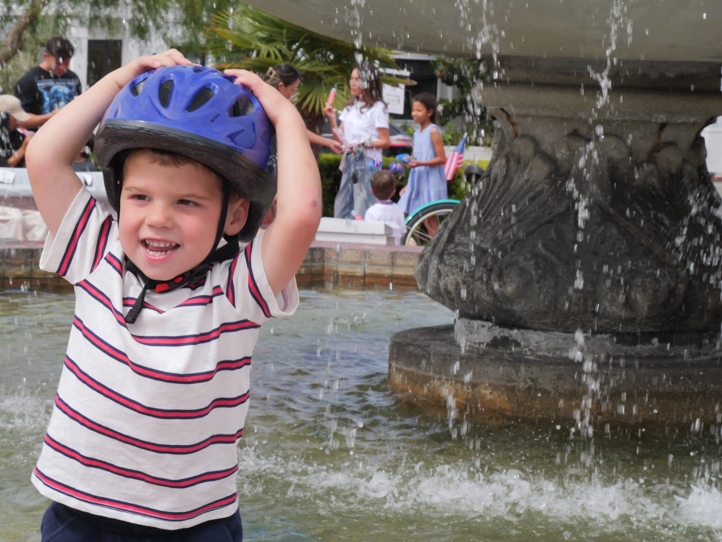 A young boy wearing a striped shirt and a blue helmet is adjusting his helmet while standing in front of a fountain with water cascading down. In the background, there are several people, including children and women, near a building decorated with p