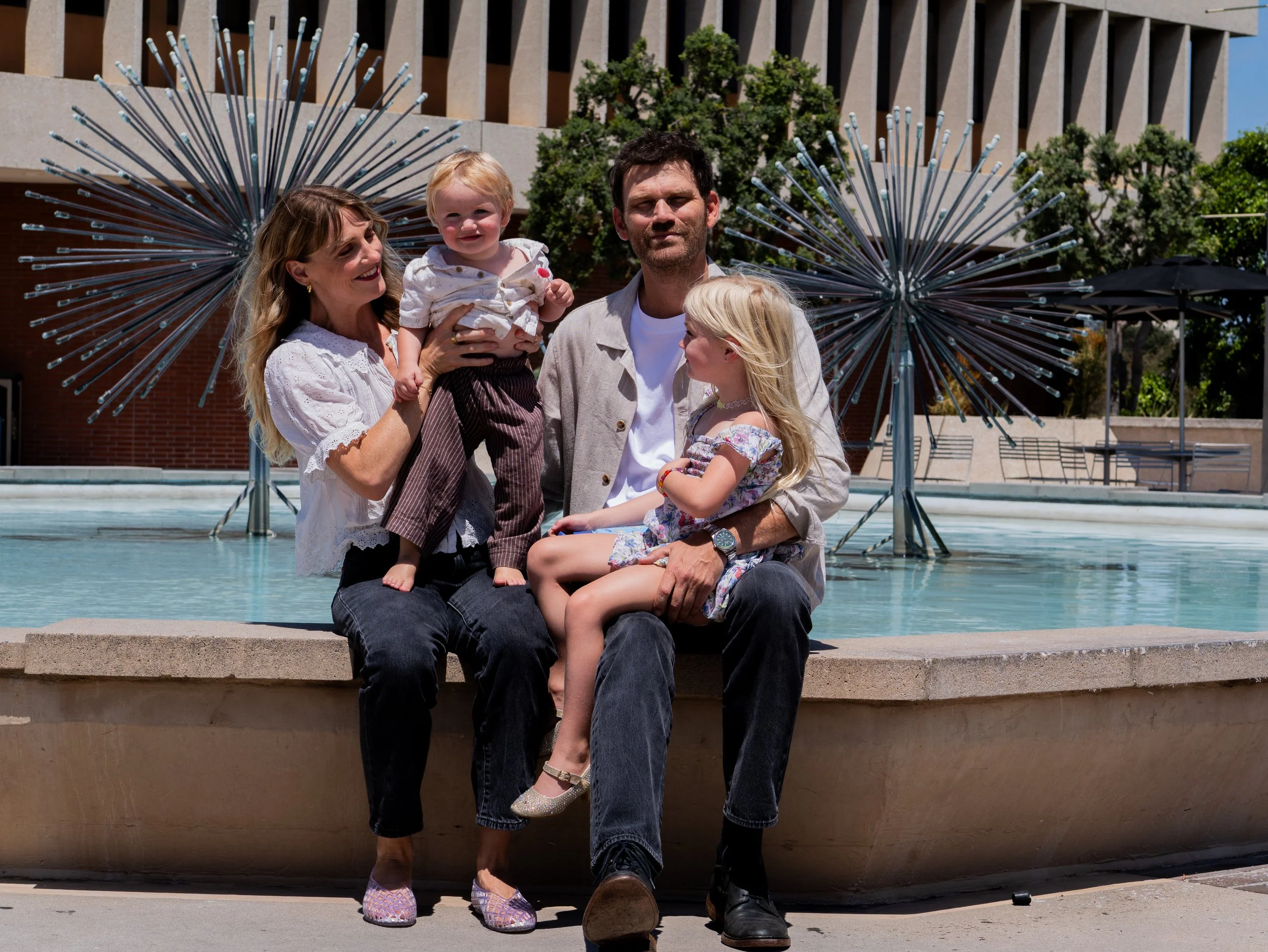 A family of four sitting near a pool with two decorative metal sculptures behind them. The family includes a woman, a man, a young girl, and a young boy, all smiling and enjoying a sunny day outside.