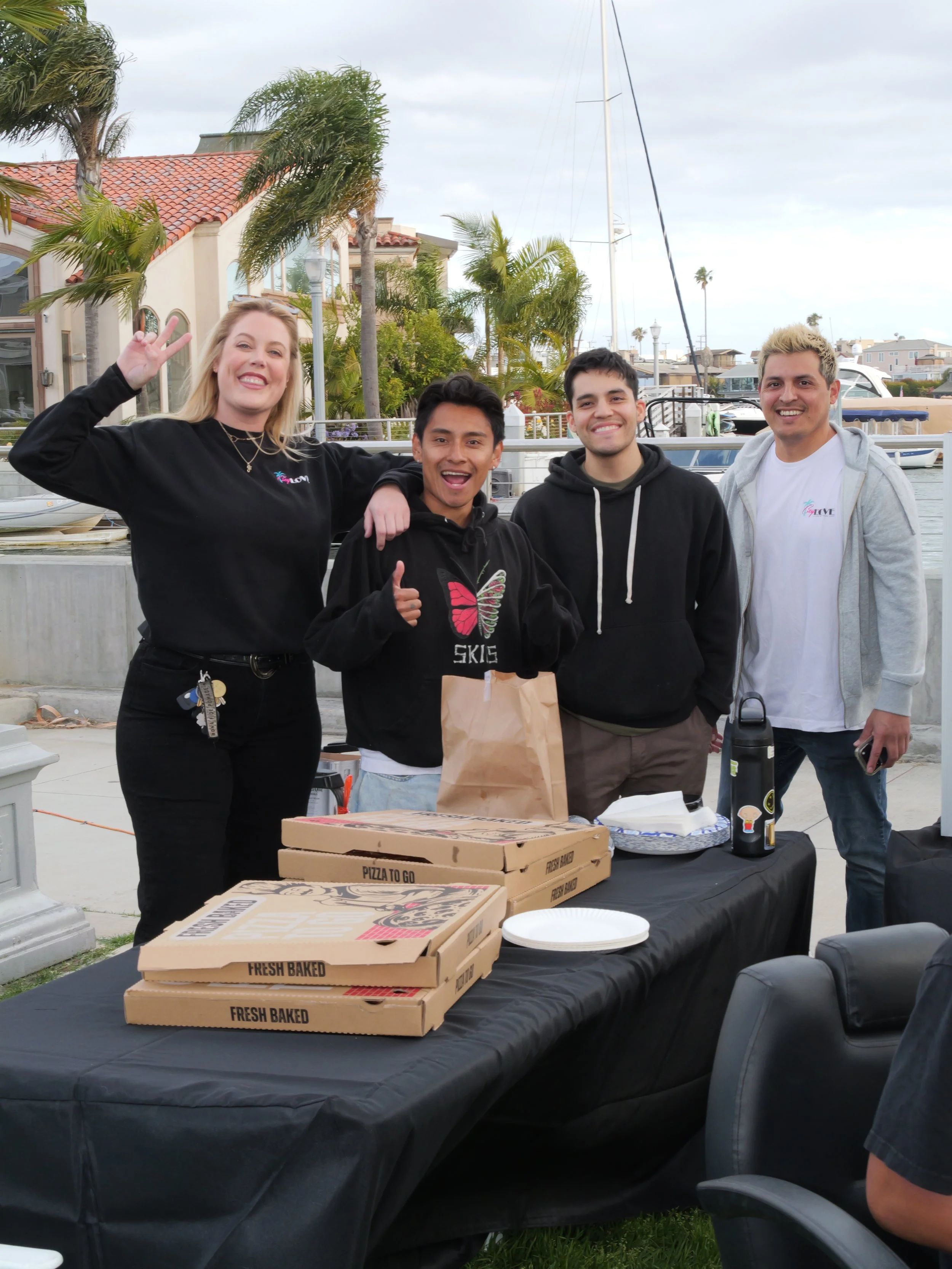 Four people smiling and posing at a gathering near a marina with boats and houses in the background. There are pizza boxes on a table in front of them.