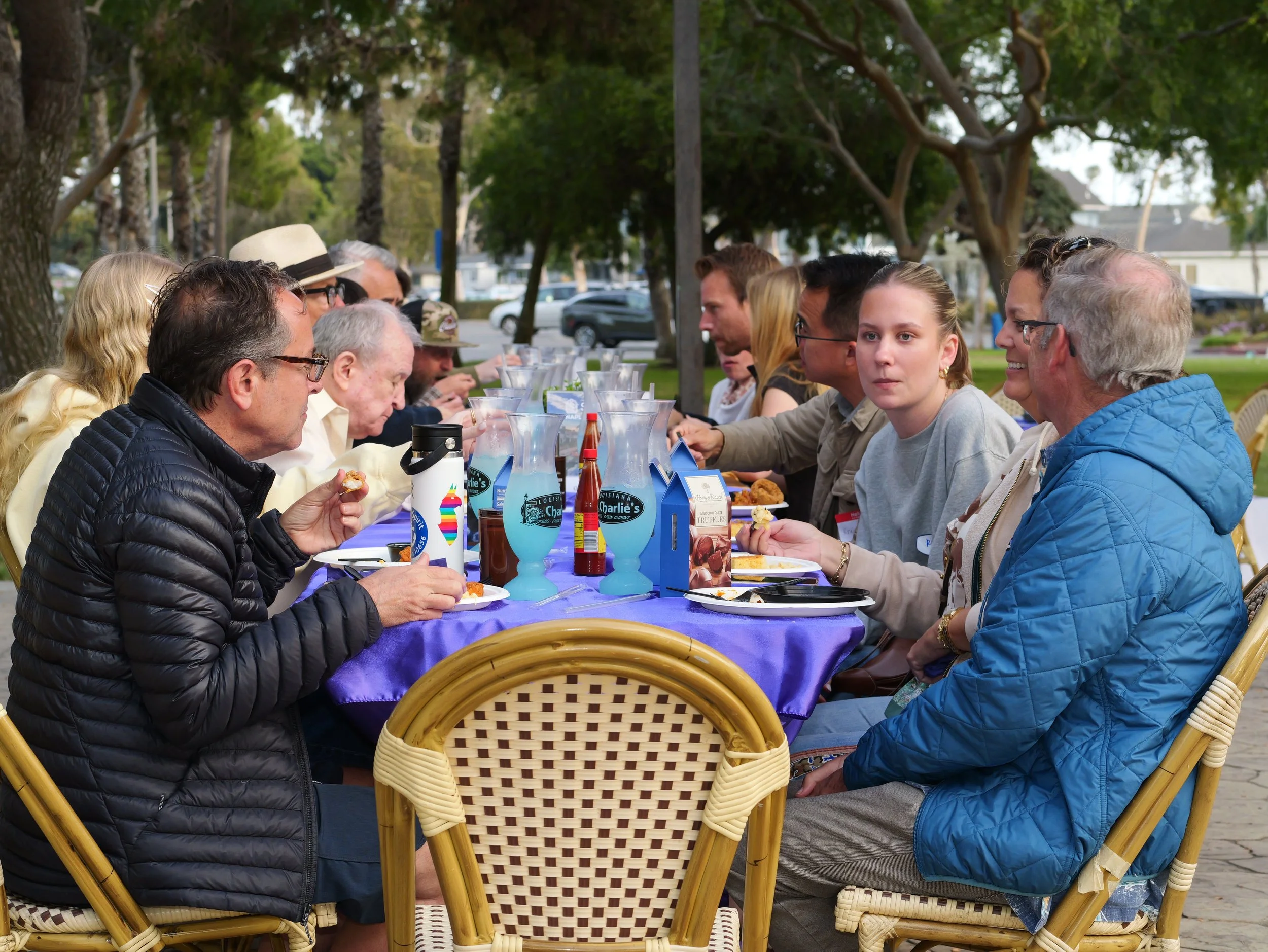 A group of people sitting at a long outdoor table under trees, enjoying food and drinks at a social gathering or event.