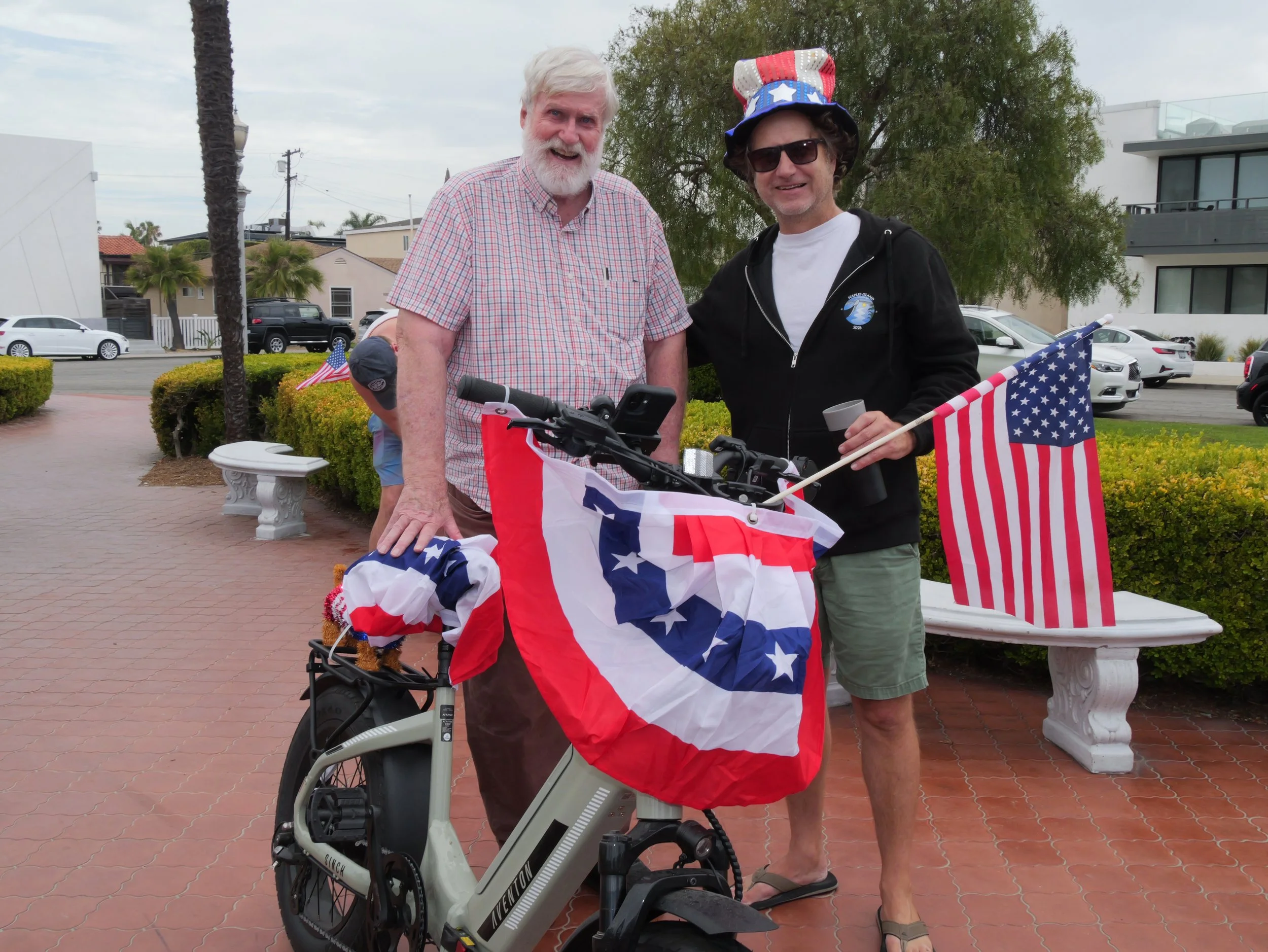 Two men standing outdoors on sidewalk, one wearing a checkered shirt with white hair and beard, the other in sunglasses, black hoodie, and patriotic hat with American flag design, holding a small flag; bicycle with American flags and decorations in f