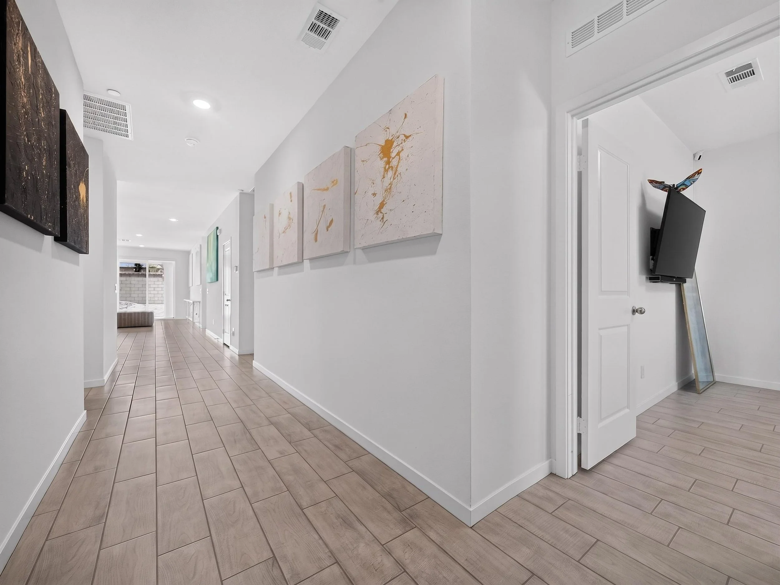 A modern, bright hallway with light-colored wood tile flooring, white walls, and artwork hanging on the wall. A doorway with a butterfly-shaped decoration on a TV is visible on the right side, and natural light enters from the back through a window.