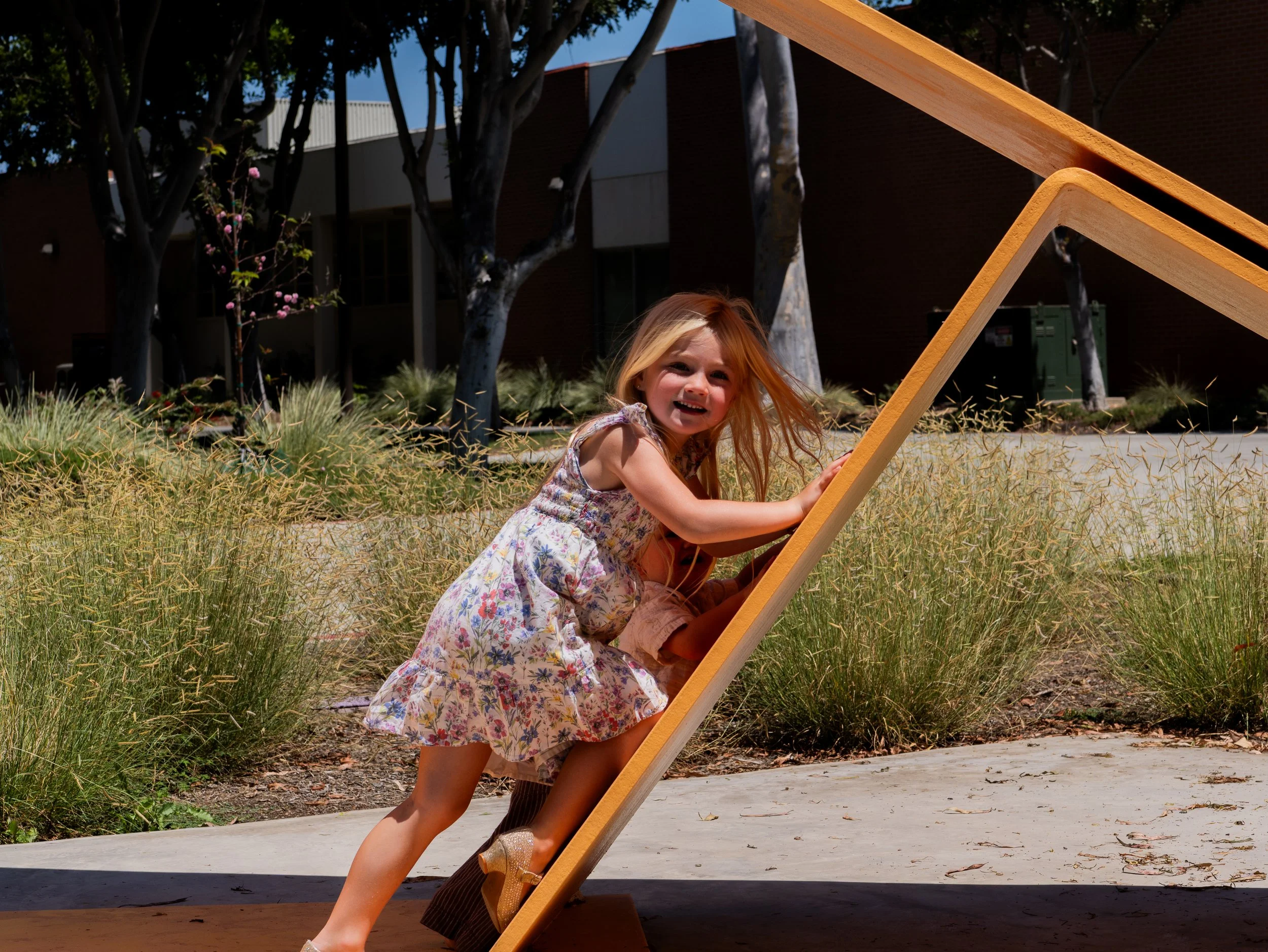 A young girl with red hair, wearing a floral dress and gold shoes, is playing outdoors on a wooden slide with a smiling and happy expression.