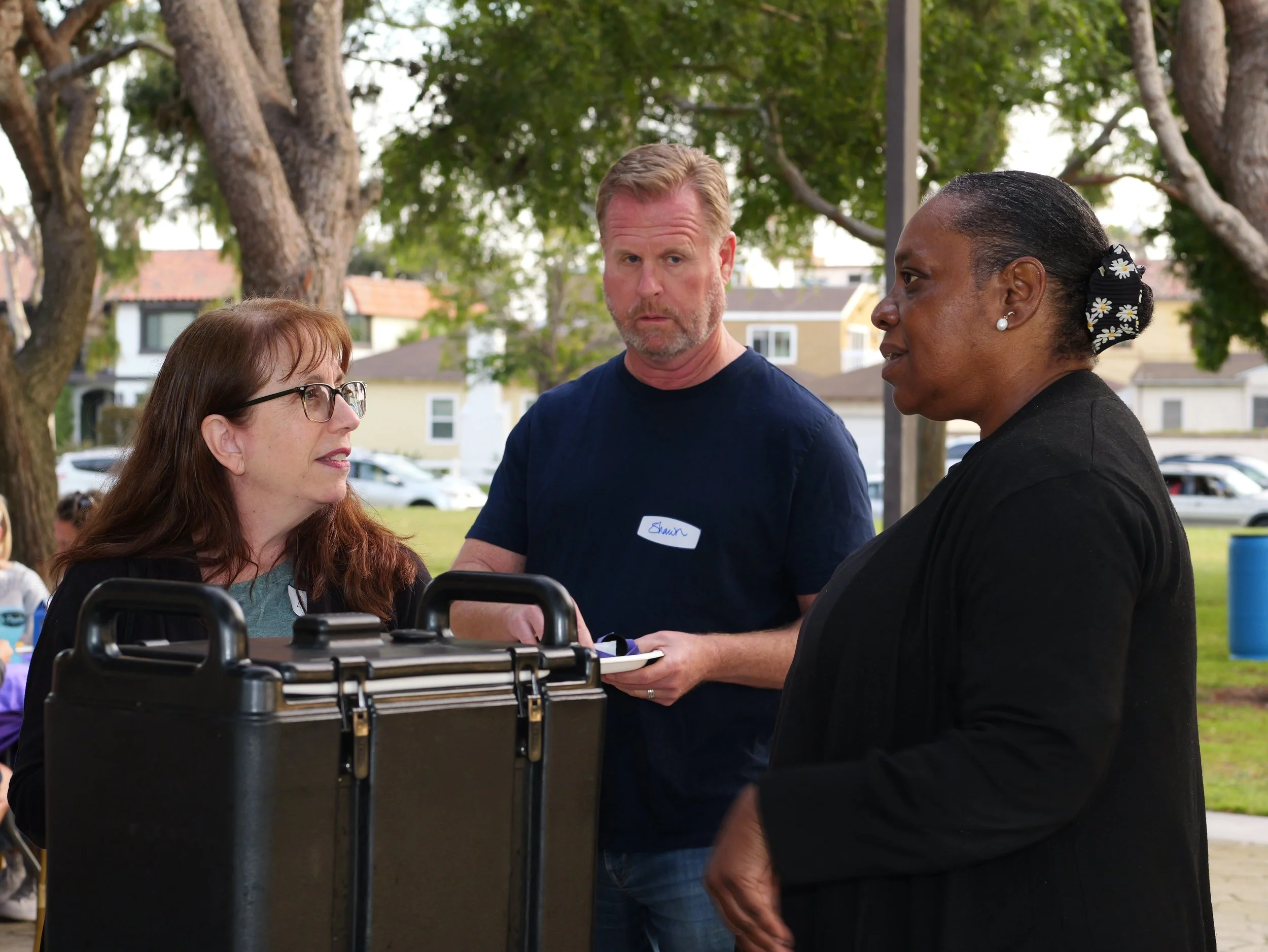 Three adults standing outdoors under trees, engaged in conversation near a black briefcase.