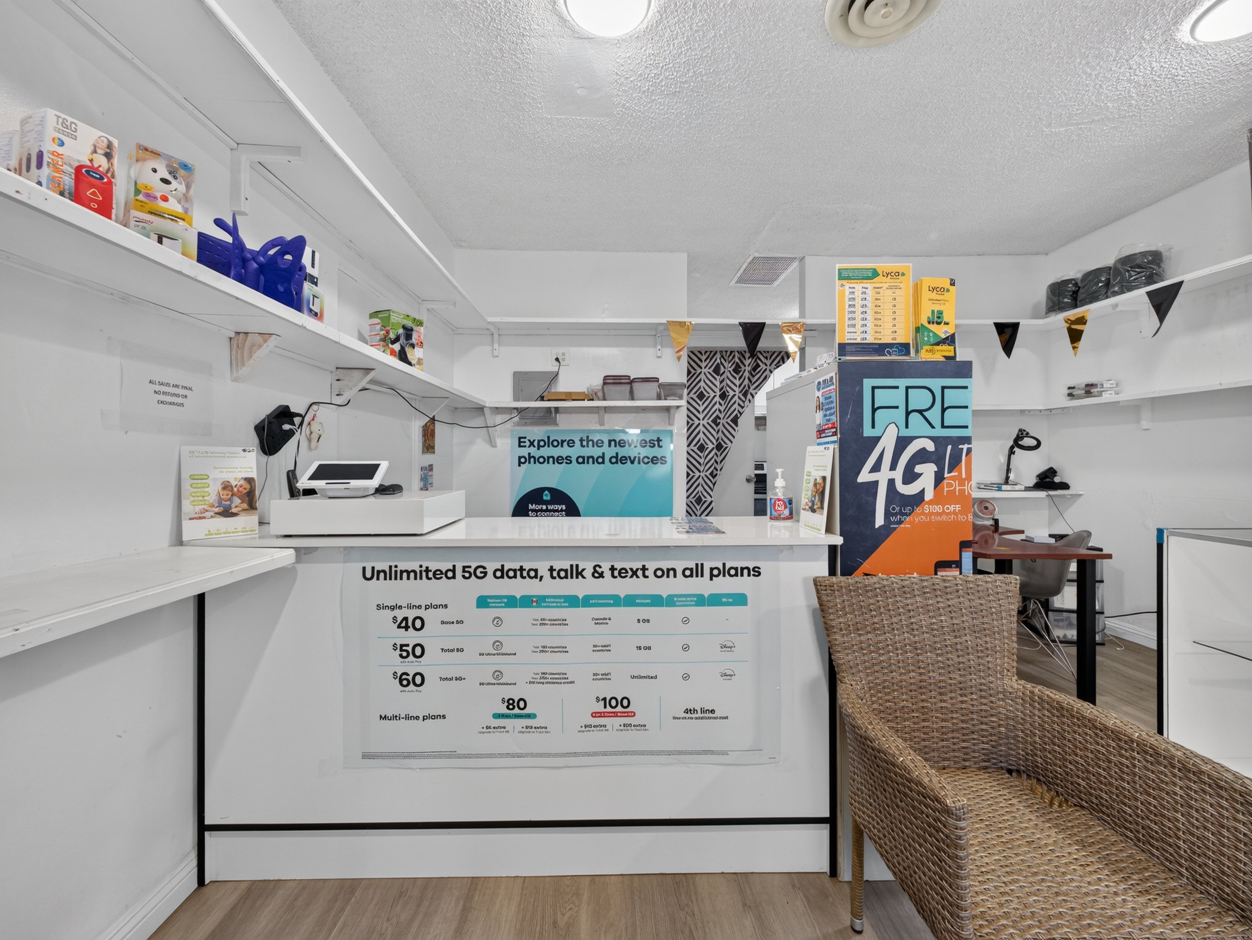 View of a retail store checkout counter with shelves displaying various small products, a computer monitor, a hand sanitizer bottle, a woven chair, and promotional signs about mobile plans.