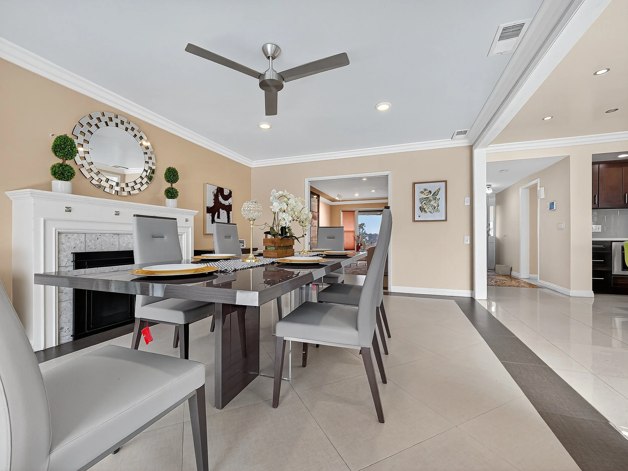 Modern dining room with a dark wood table set for four, white upholstered chairs, a white fireplace mantle with decorative greenery, a round mirror, and a flower arrangement in the center. The ceiling has a silver ceiling fan and recessed lighting.