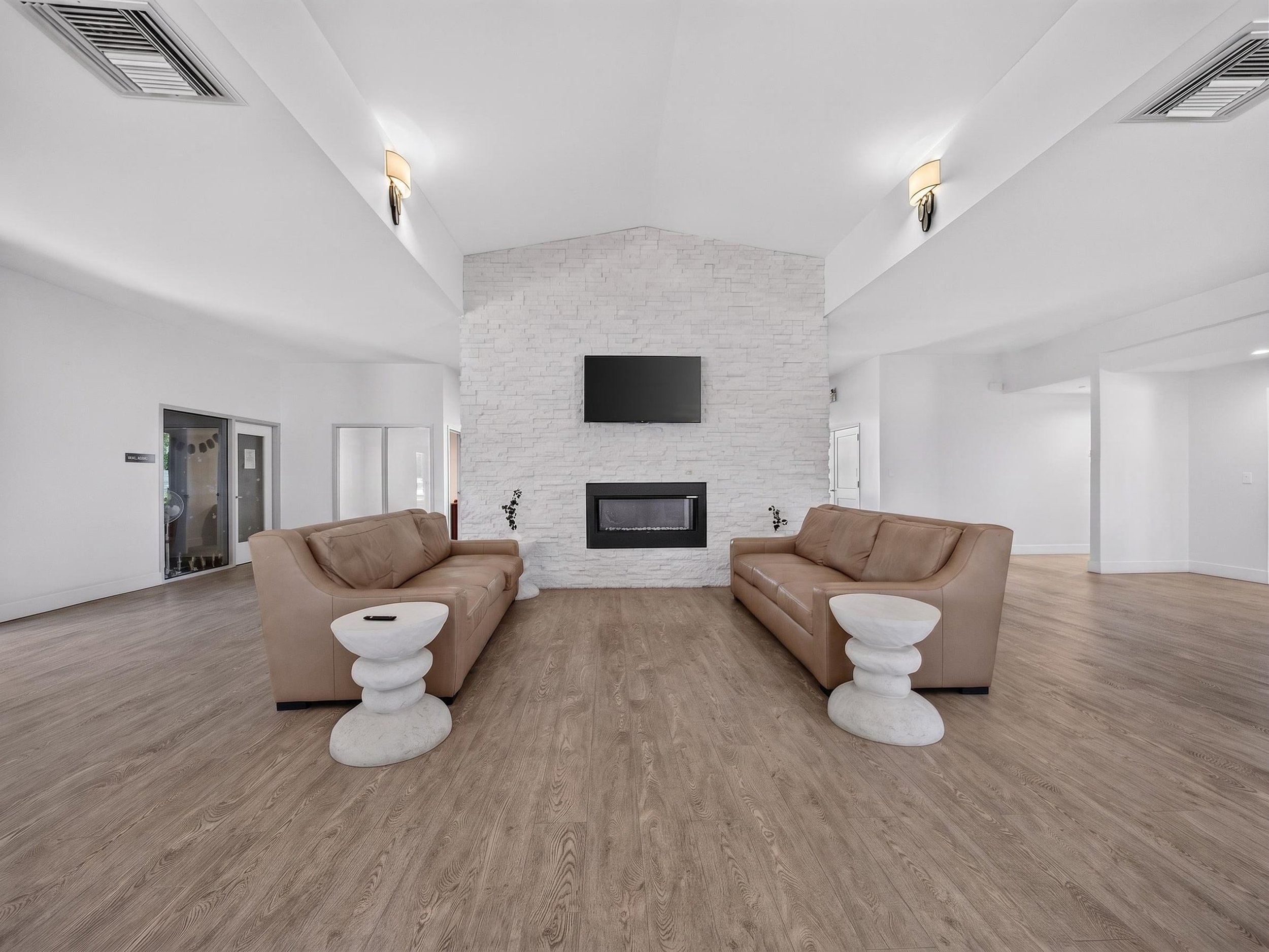 Modern living room with beige sofas, white side tables, a white brick fireplace, flat-screen TV, and light-colored wooden flooring.