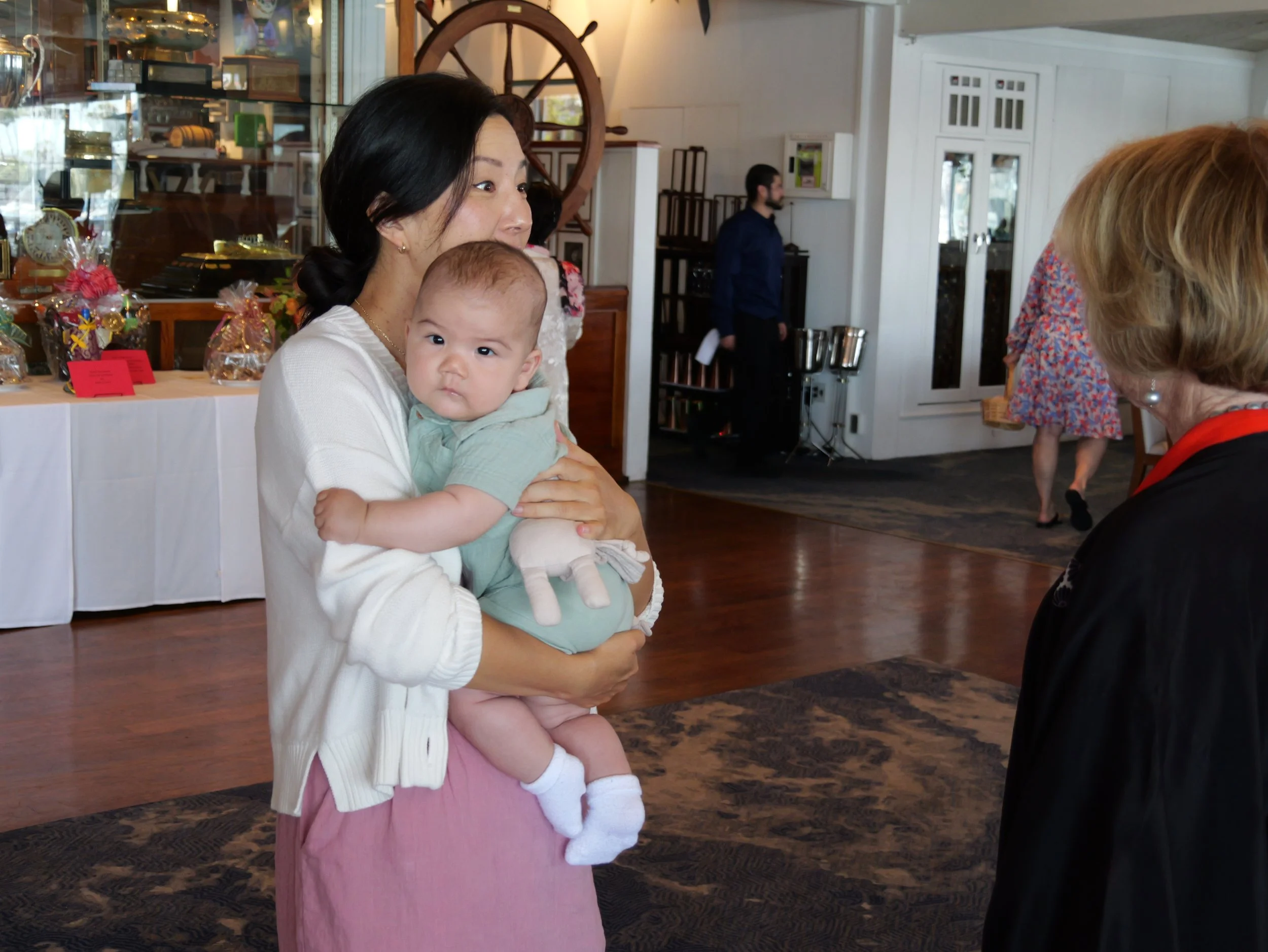 A woman with dark hair holding a baby in a light green outfit, at an indoor event. The woman is smiling and looking to her right, while the baby looks at the camera. There are others in the background, including a person with blonde hair and a woman 